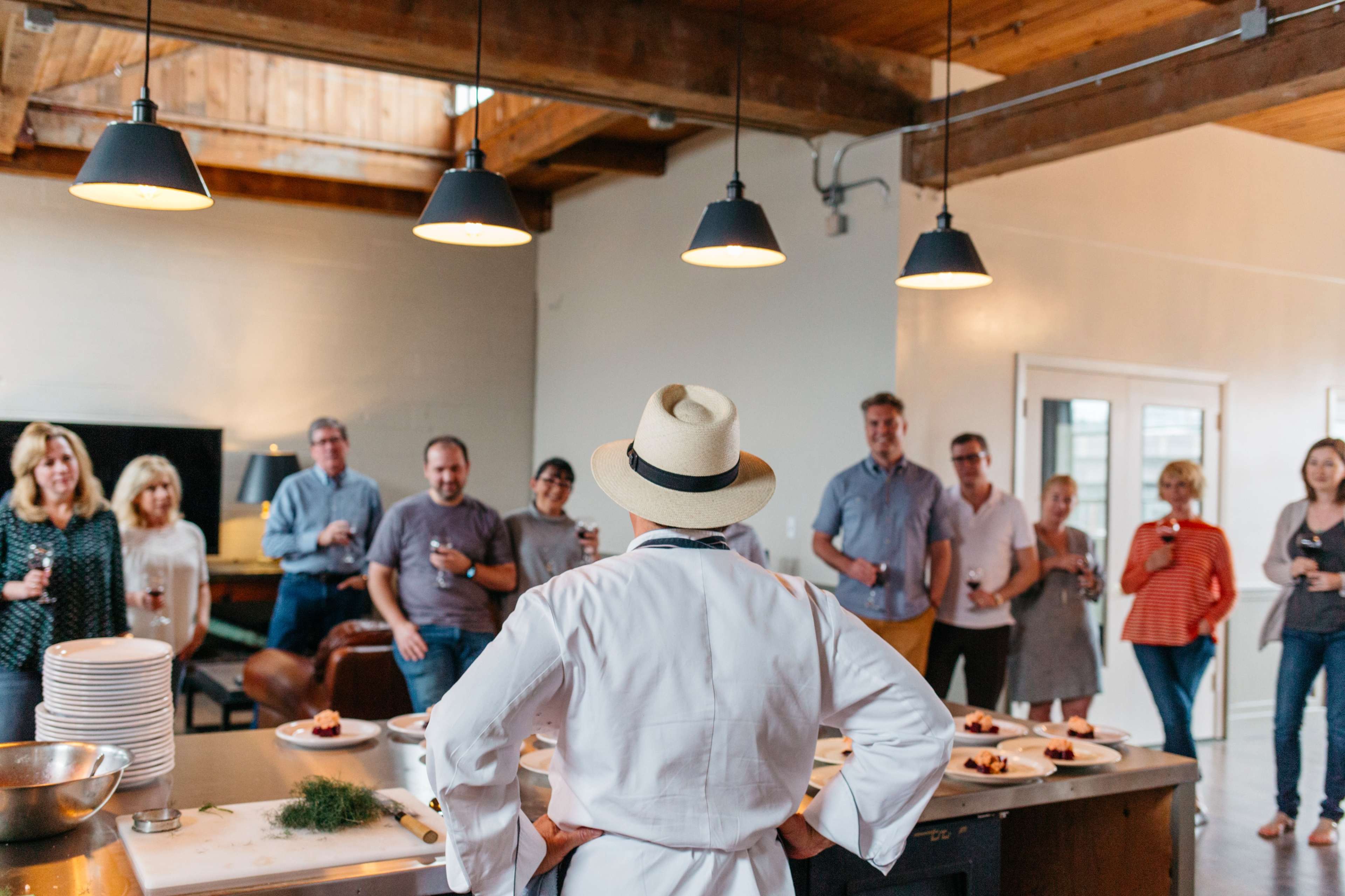 A group of people in a contemporary kitchen watches a chef presenting a plated dish.