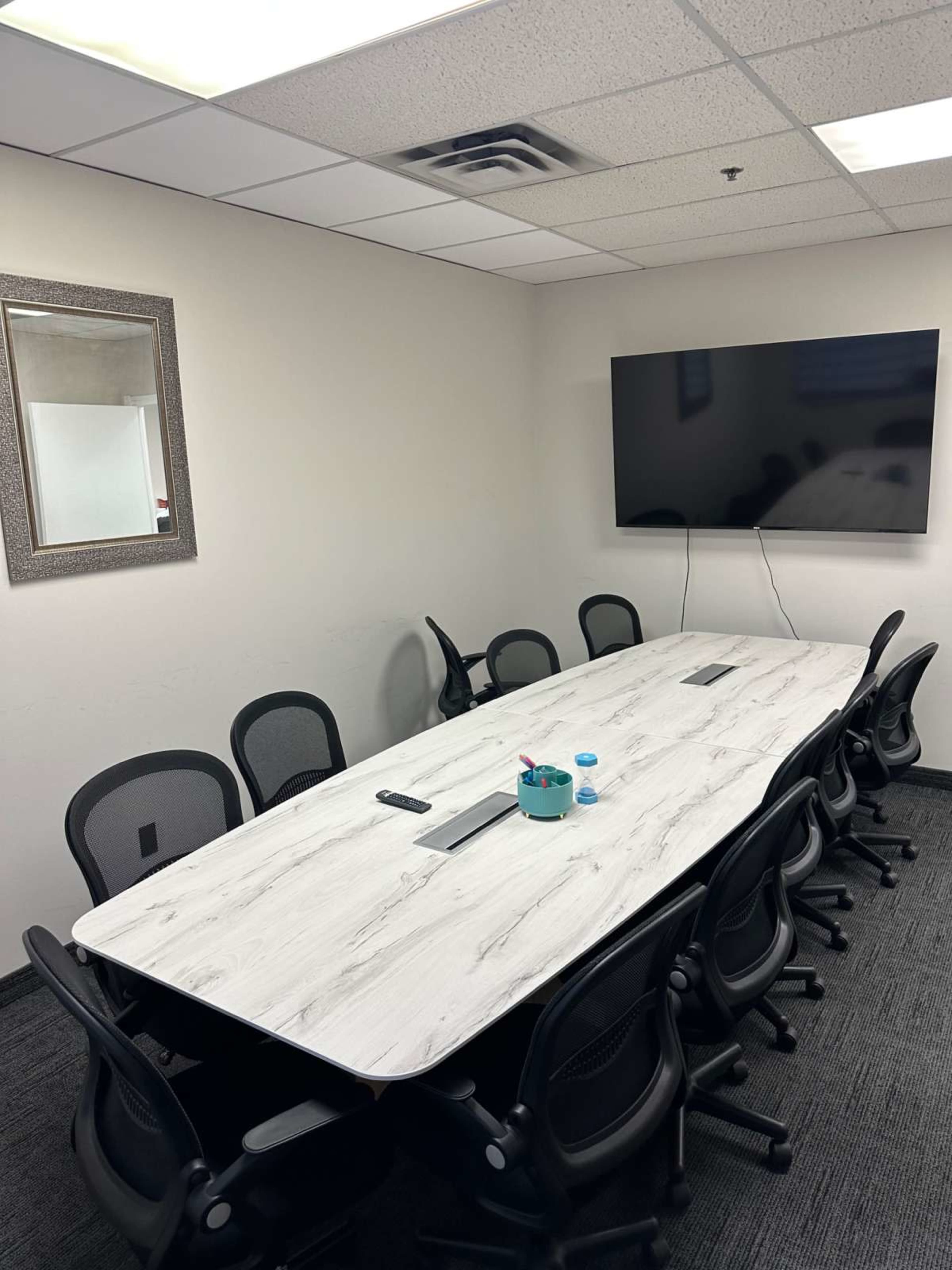 The image shows a conference room with a long, rectangular table surrounded by office chairs, a large wall-mounted TV, and a mirror on the wall.