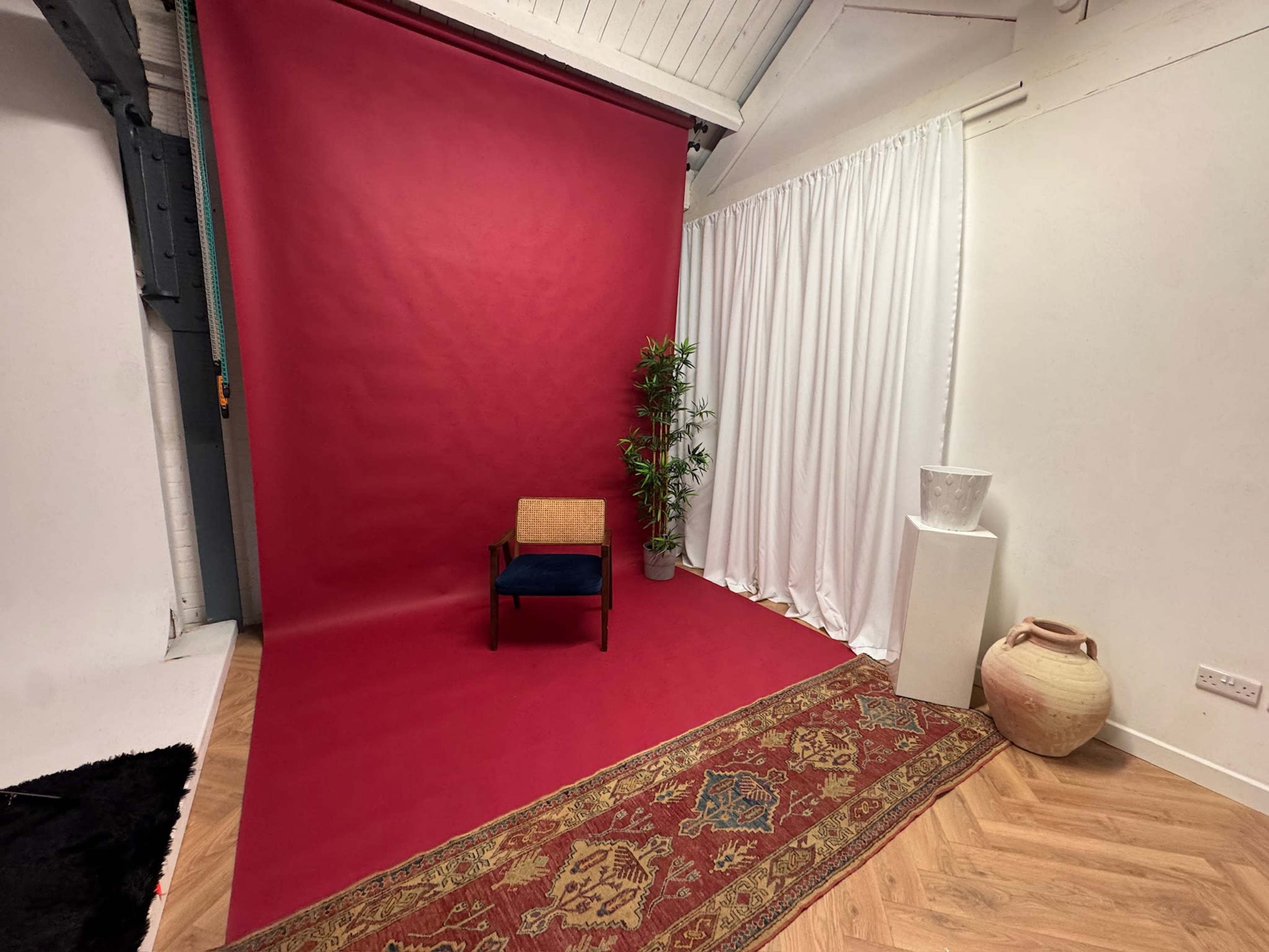 The image shows a studio space featuring a vibrant red backdrop, a small wooden chair, a potted plant, a white draped curtain, and decorative items on a pedestal.