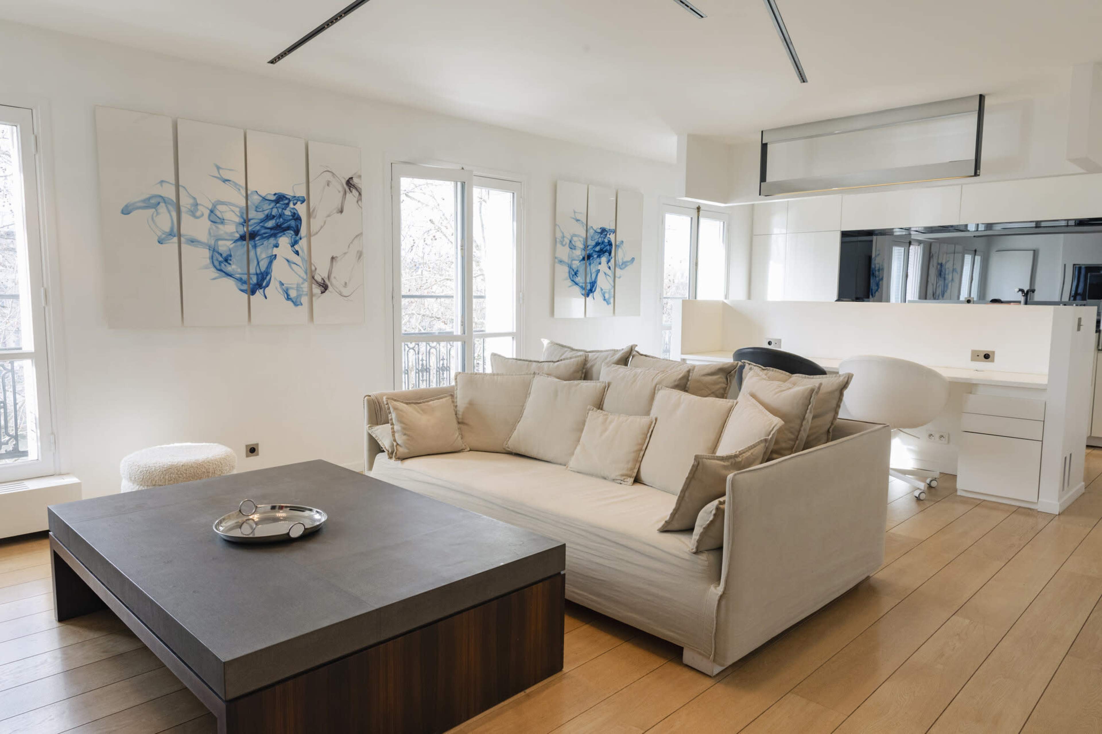 The image shows a modern living room with a large beige sofa adorned with multiple cushions, a dark coffee table, and abstract artwork on the walls, all set in a bright space with large windows.
