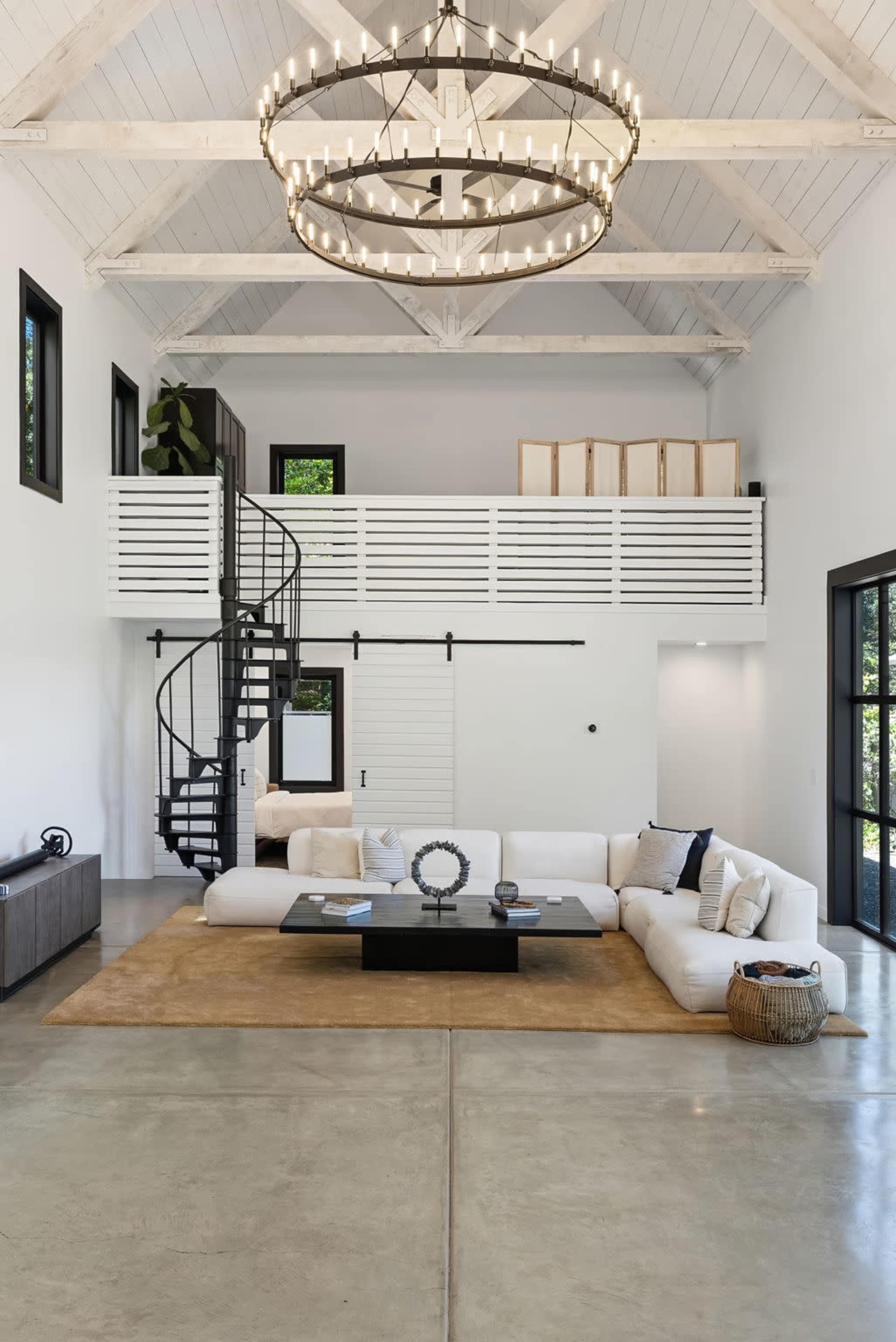 The image depicts a spacious, modern living room with high ceilings, a spiral staircase, and minimalist furniture centered around a coffee table on a large area rug.
