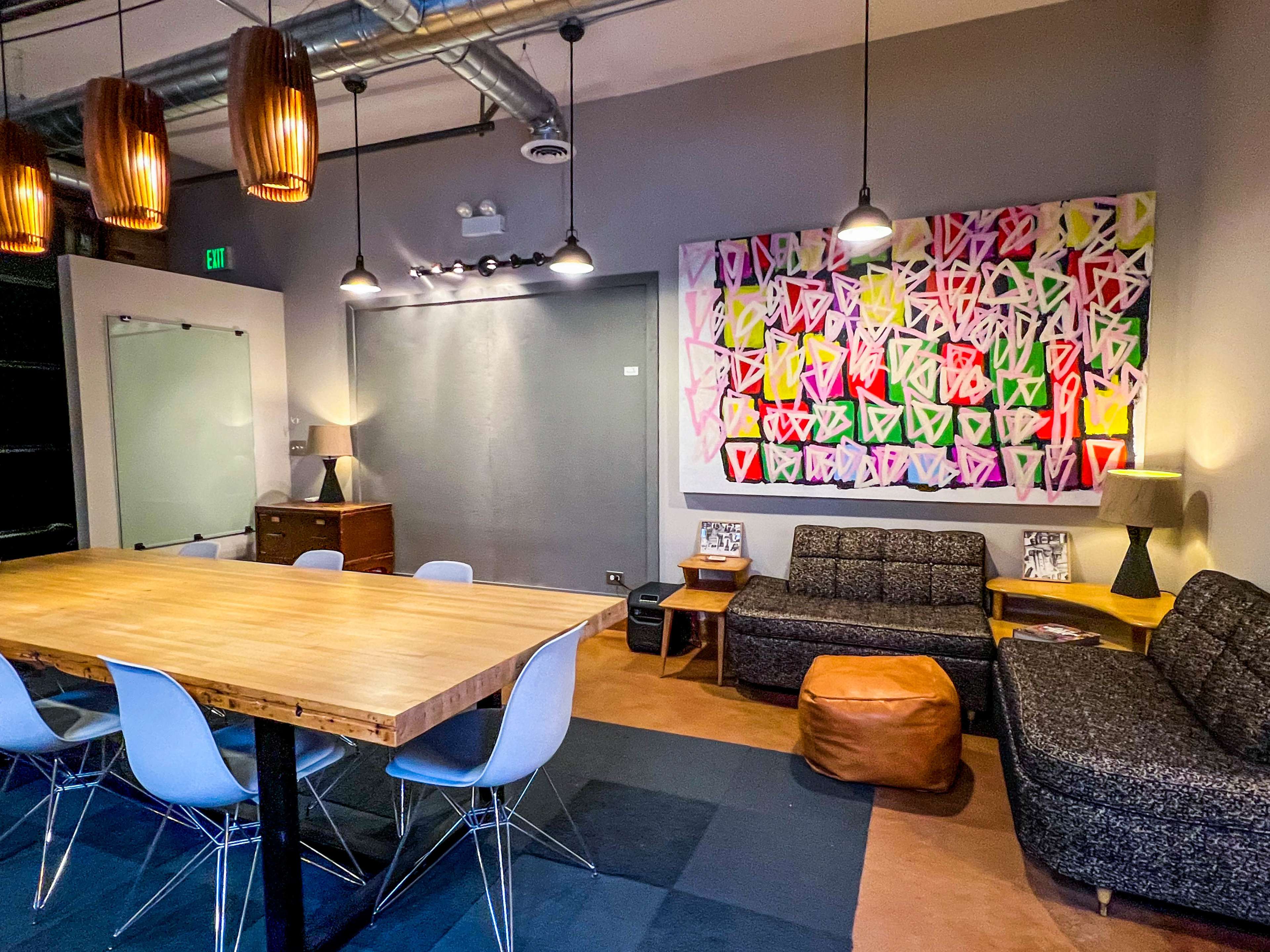 The image shows a modern lounge area with a large wooden table, several chairs, two sofas, a pouf, and a colorful abstract painting on the wall.