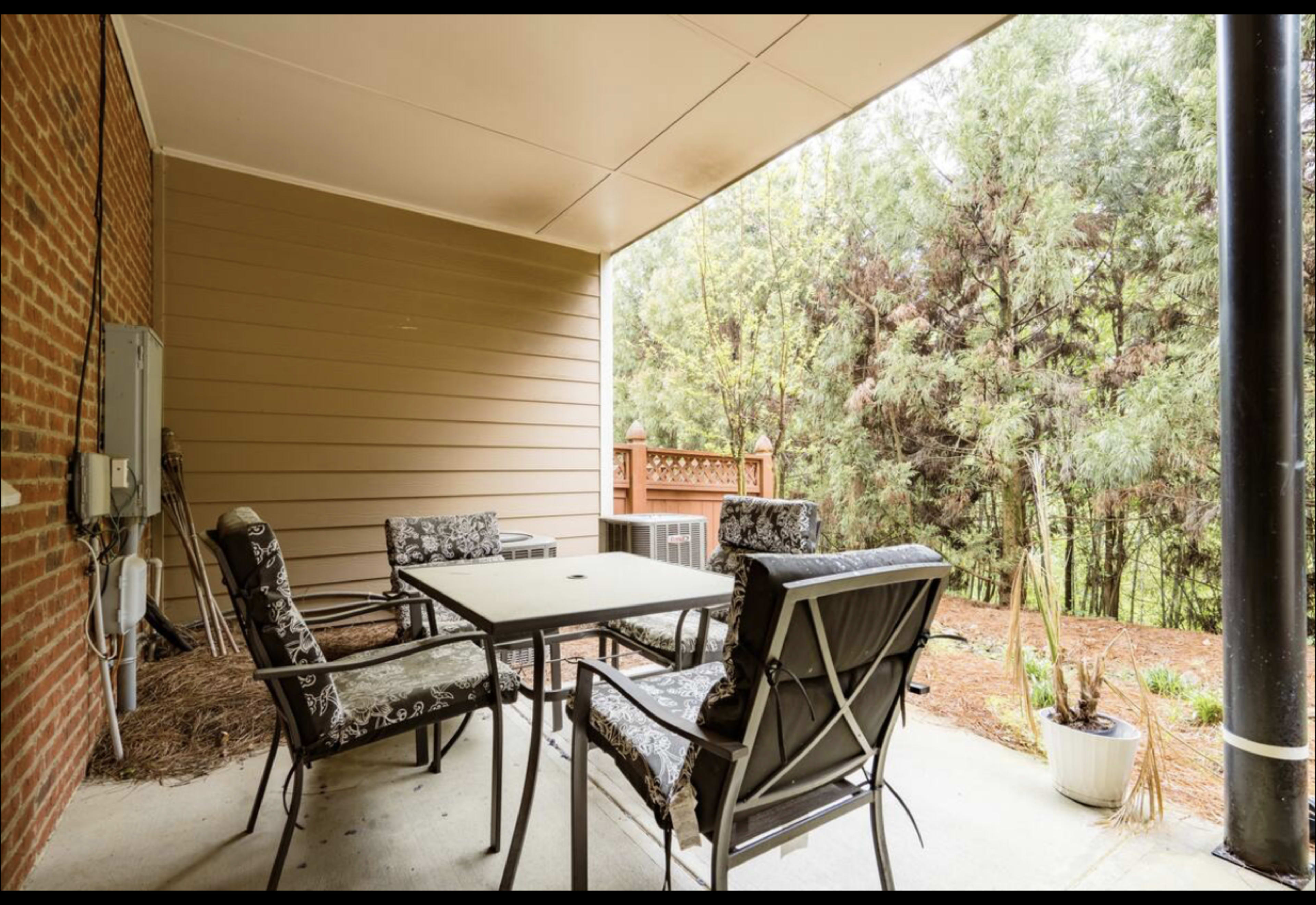 A covered patio area with a table and four chairs surrounded by greenery.