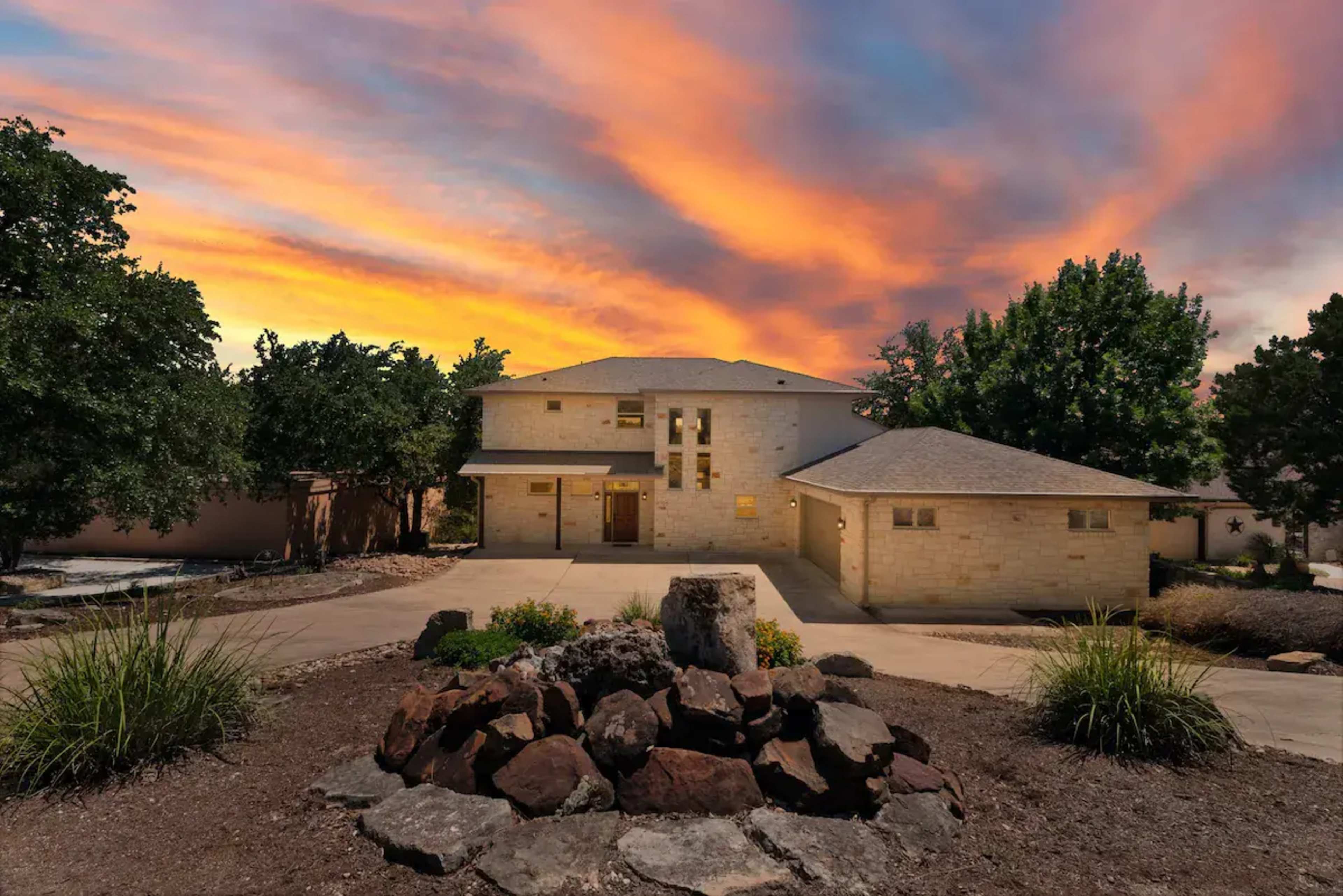 A modern stone house is illuminated against a vibrant sunset, surrounded by landscaping and rock features.