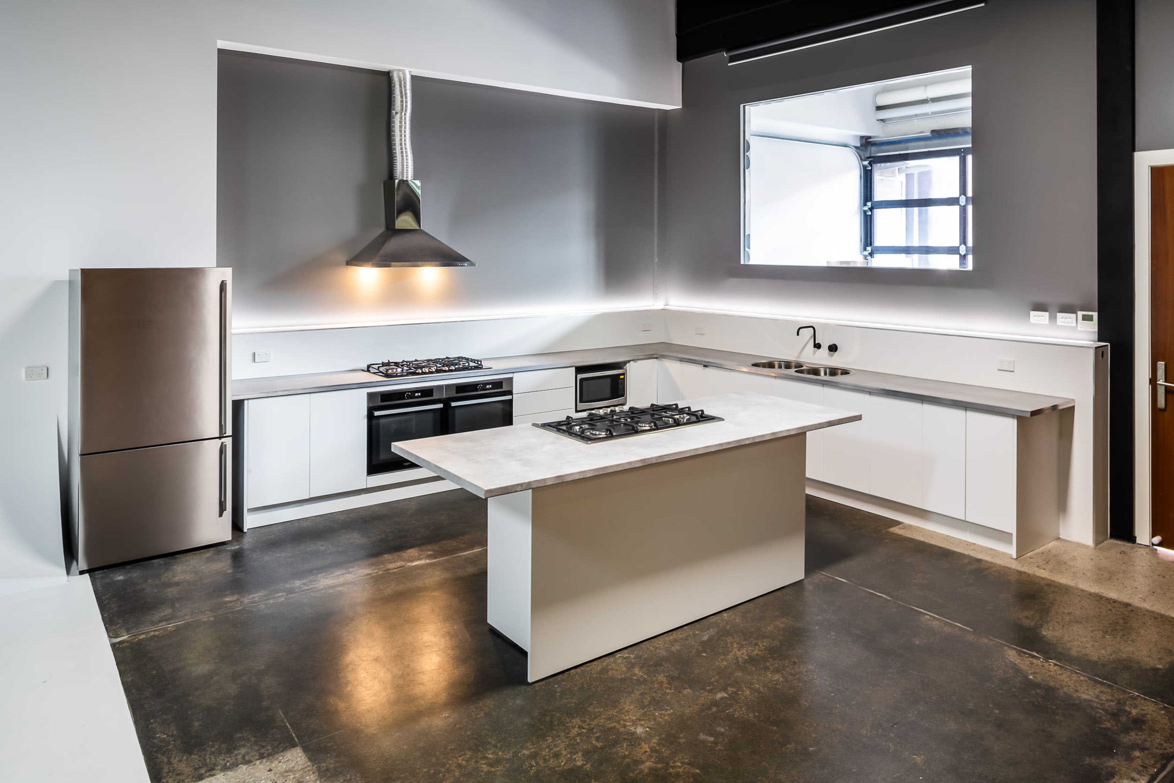 A modern kitchen features a large island with a gas cooktop, stainless steel appliances, and minimalist cabinetry against a gray wall.