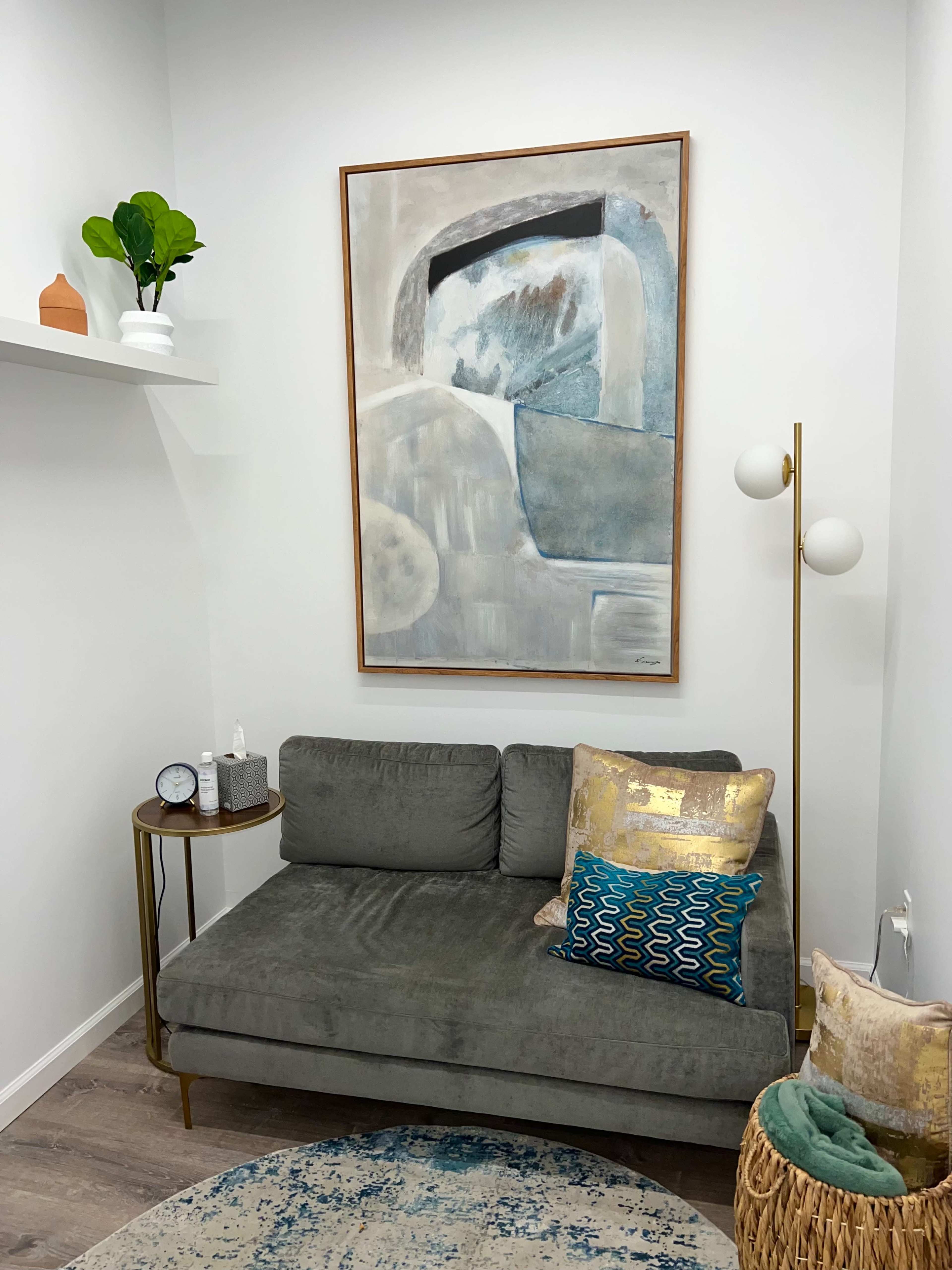 A modern living space featuring a gray sofa, a round side table with a clock, a floor lamp, and an abstract painting mounted on the white wall above the couch.