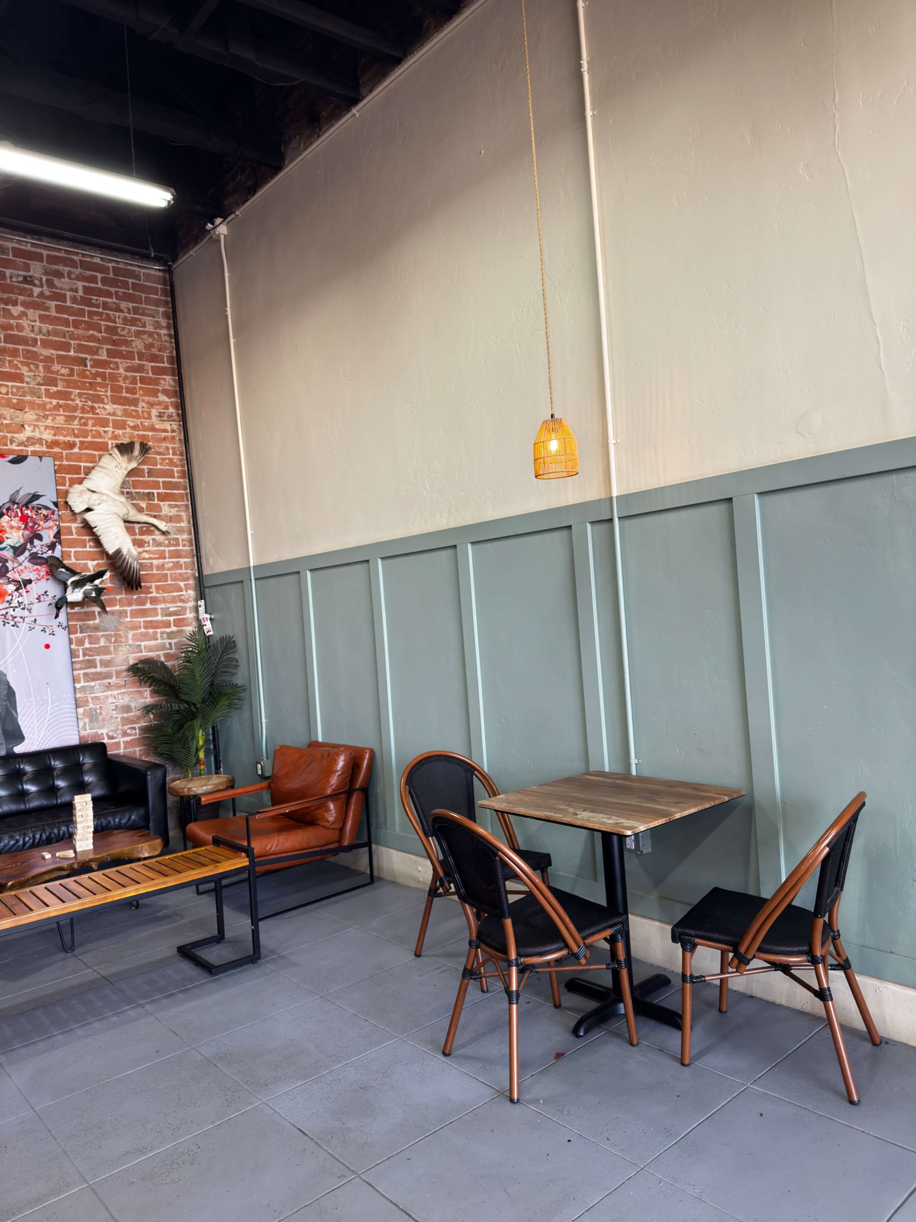 The image shows a cafe interior with a combination of brick and painted walls, featuring a mix of seating options, including a leather chair and a small table with chairs.