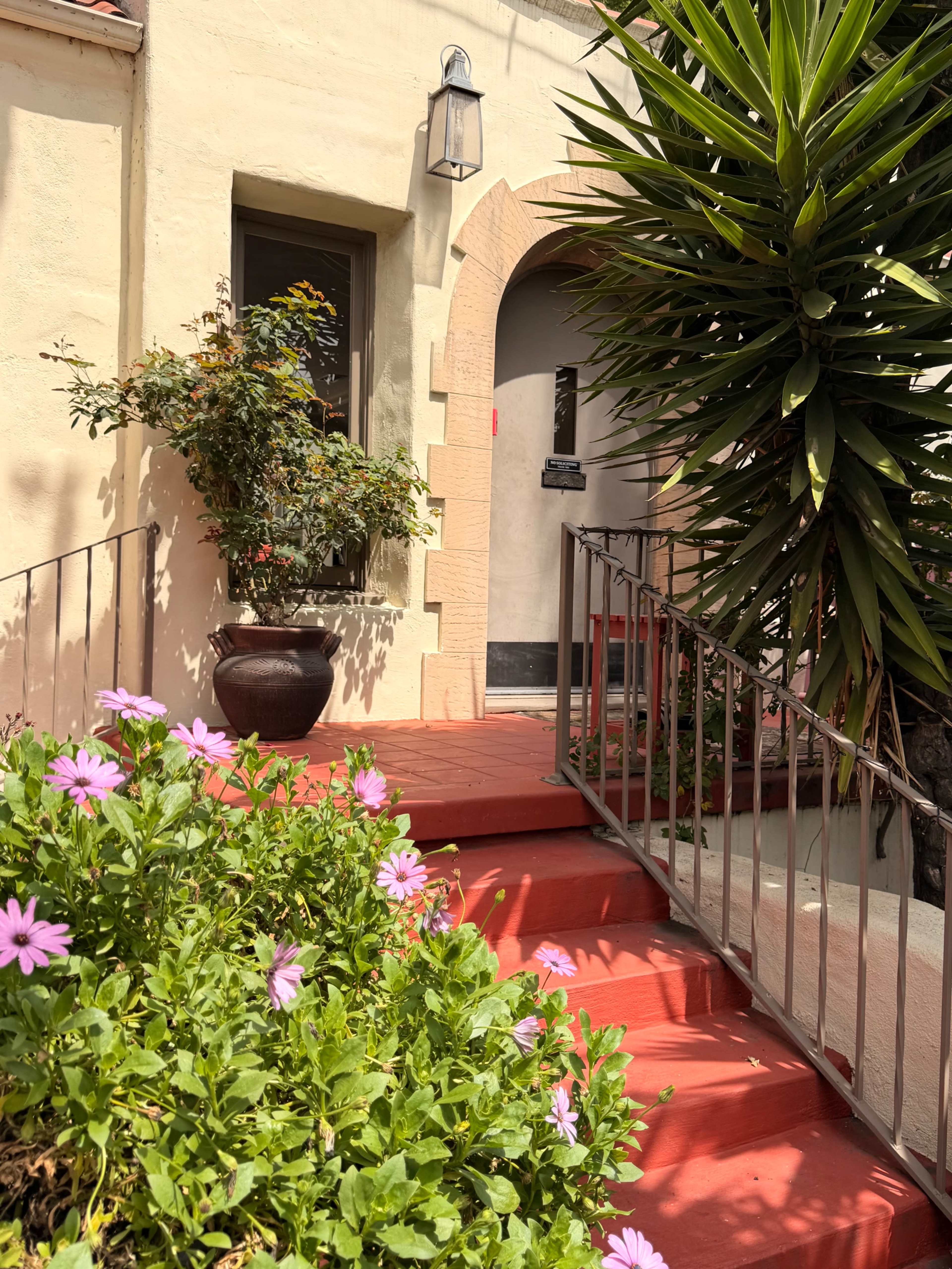 A red pathway leads up to a doorway framed by a large green plant and blooming flowers in a pot.