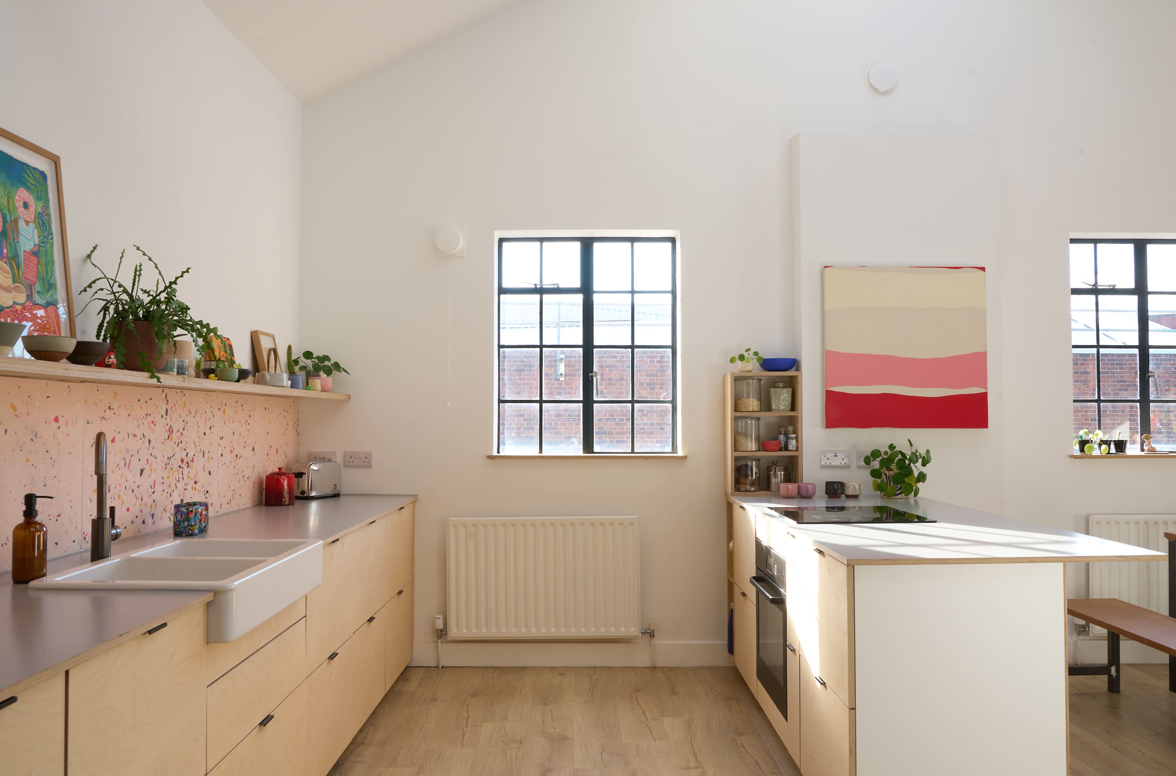 The image shows a modern kitchen with wooden cabinetry, a large window, and an open layout featuring a sink and a stove.