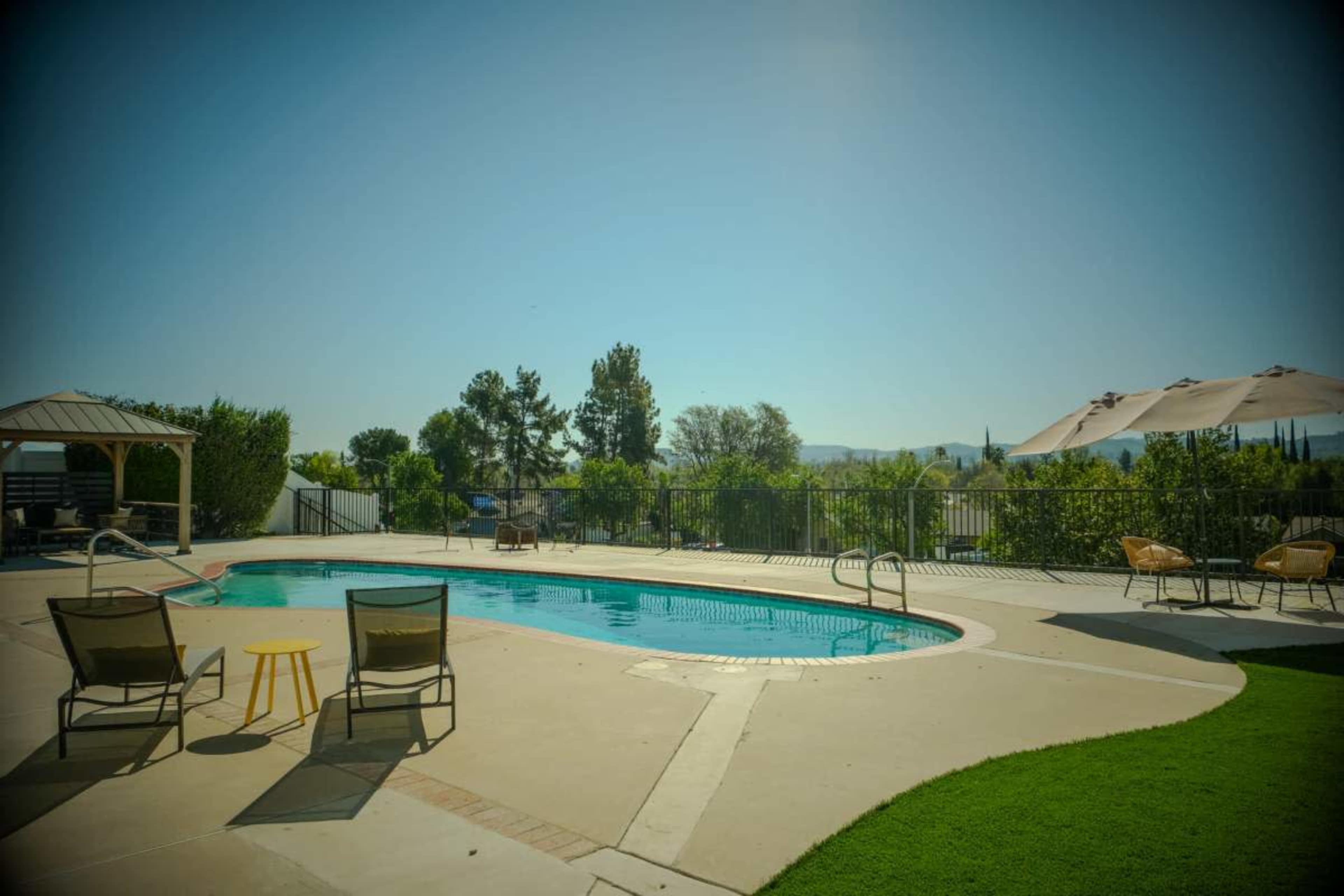 The image shows a clear blue swimming pool situated in a backyard with lounge chairs and a shaded area nearby.