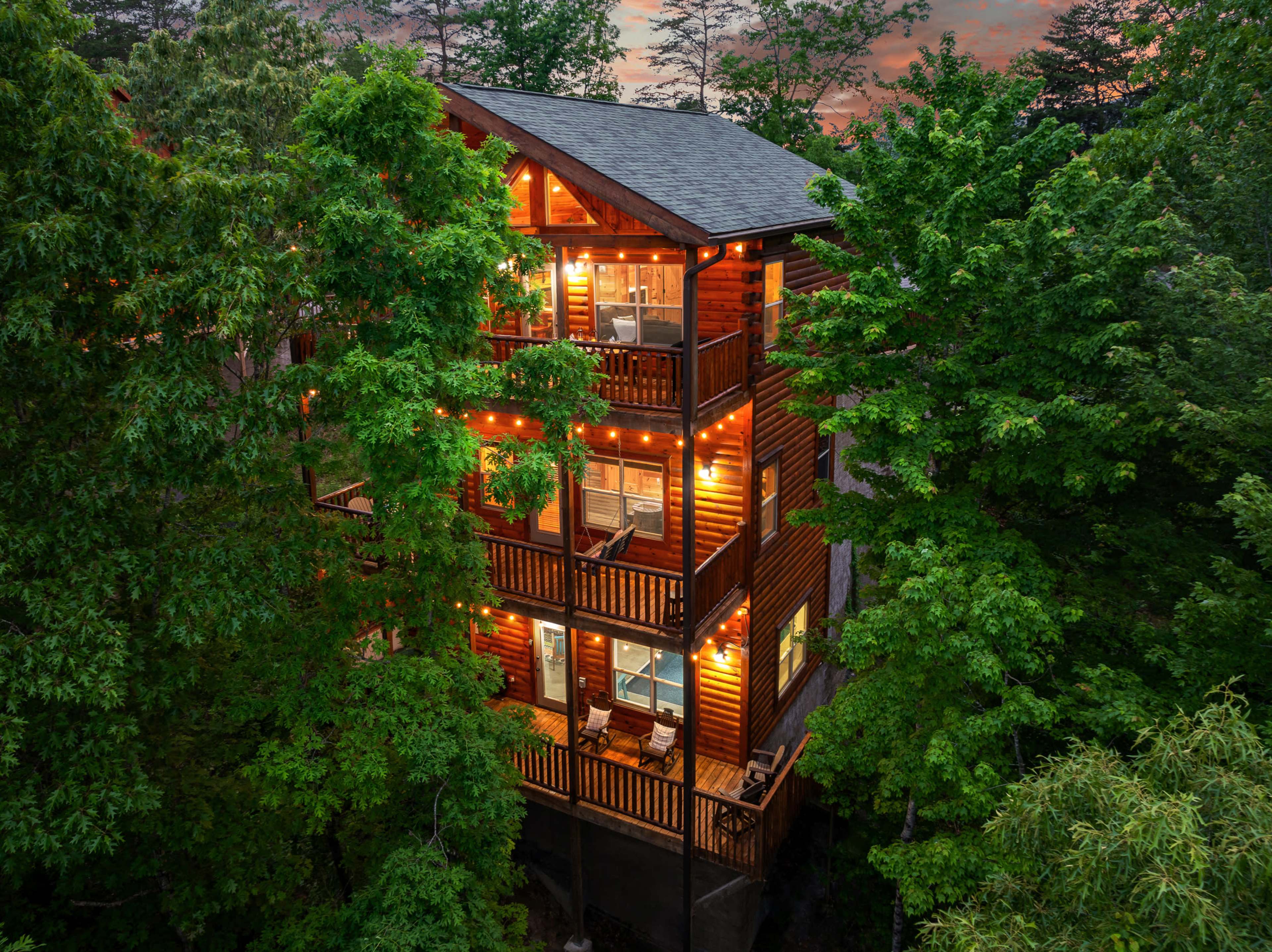 A three-story wooden cabin is surrounded by lush greenery, with lights illuminating its balconies at dusk.