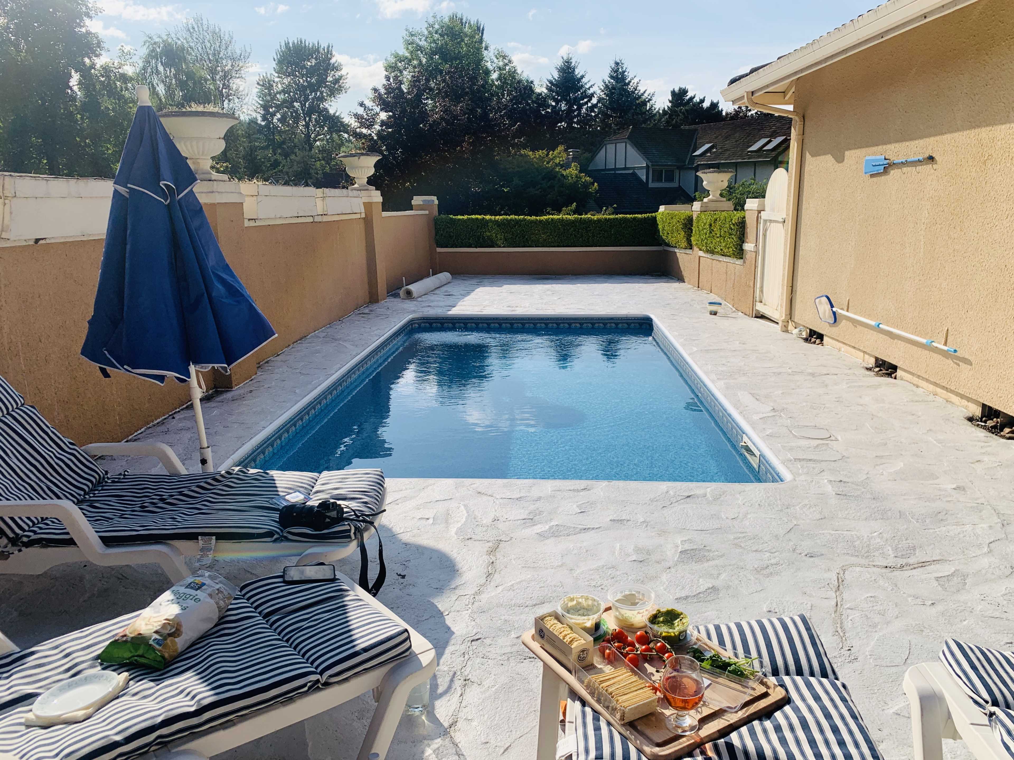 A backyard pool area features a blue pool surrounded by lounge chairs and a table with various food items.
