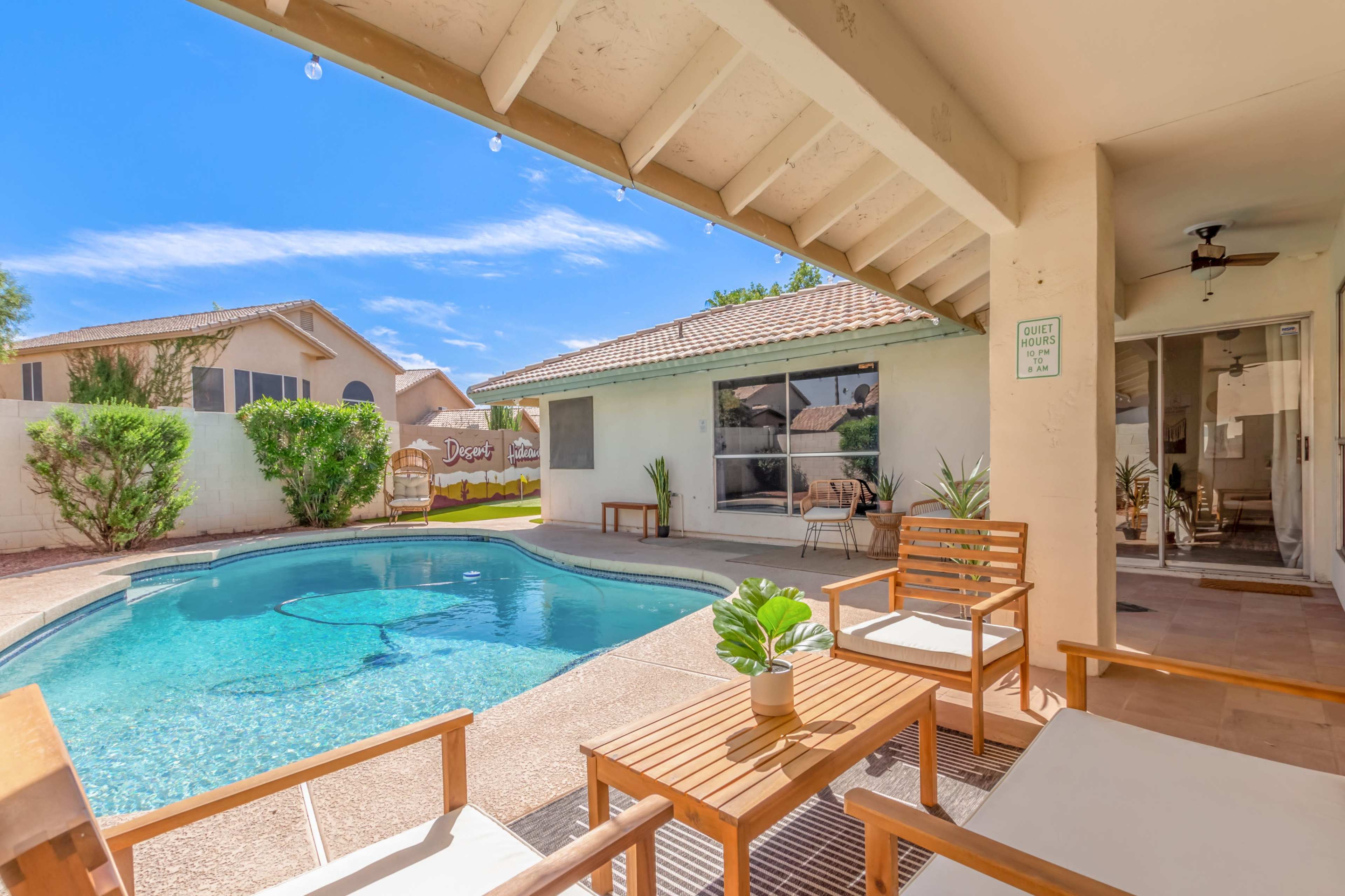 A patio area with wooden furniture overlooks a swimming pool surrounded by a landscaped yard and neighboring houses.