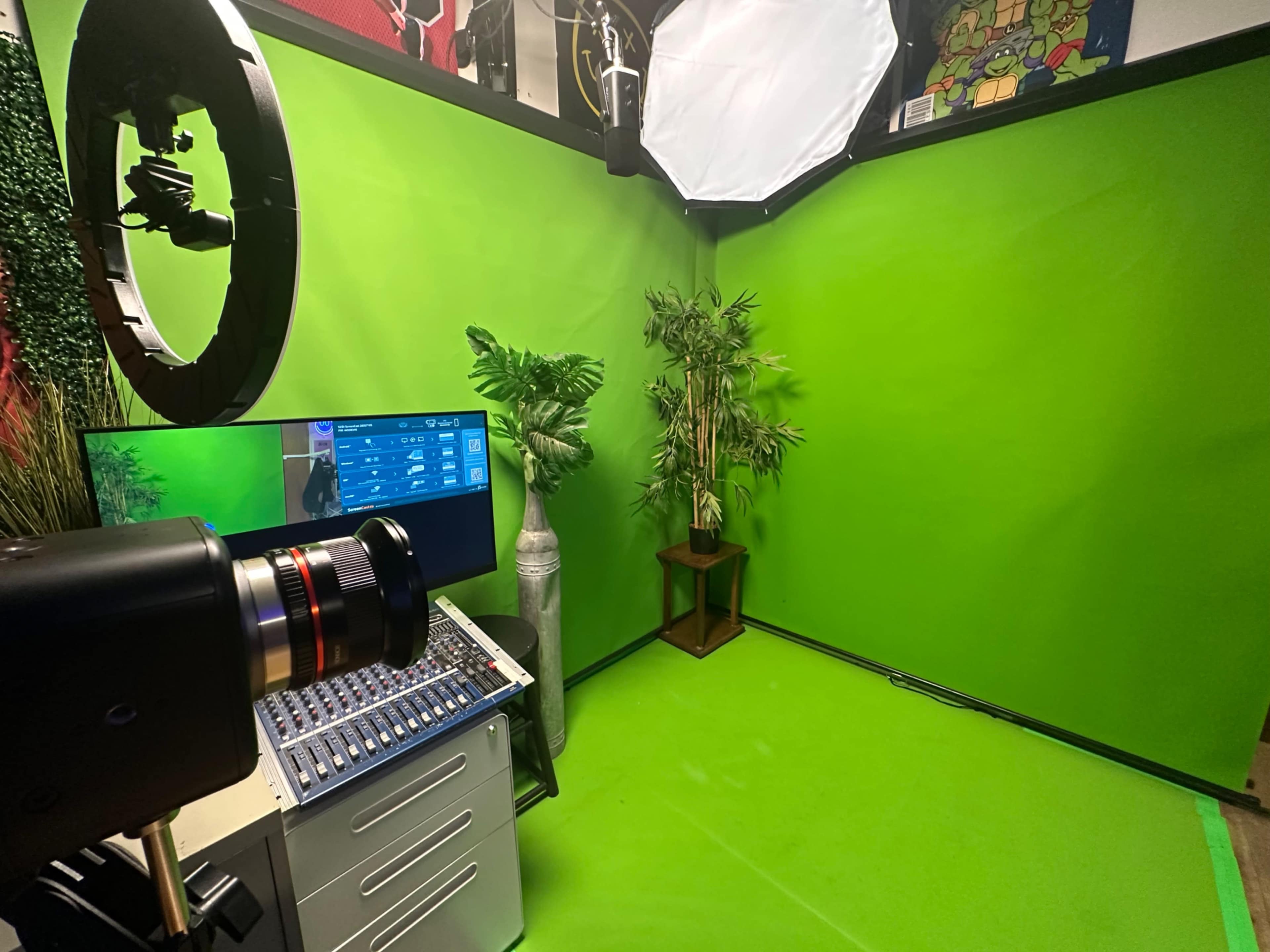 The image shows a green screen studio with a camera, a video mixer, and a potted plant, surrounded by green walls.