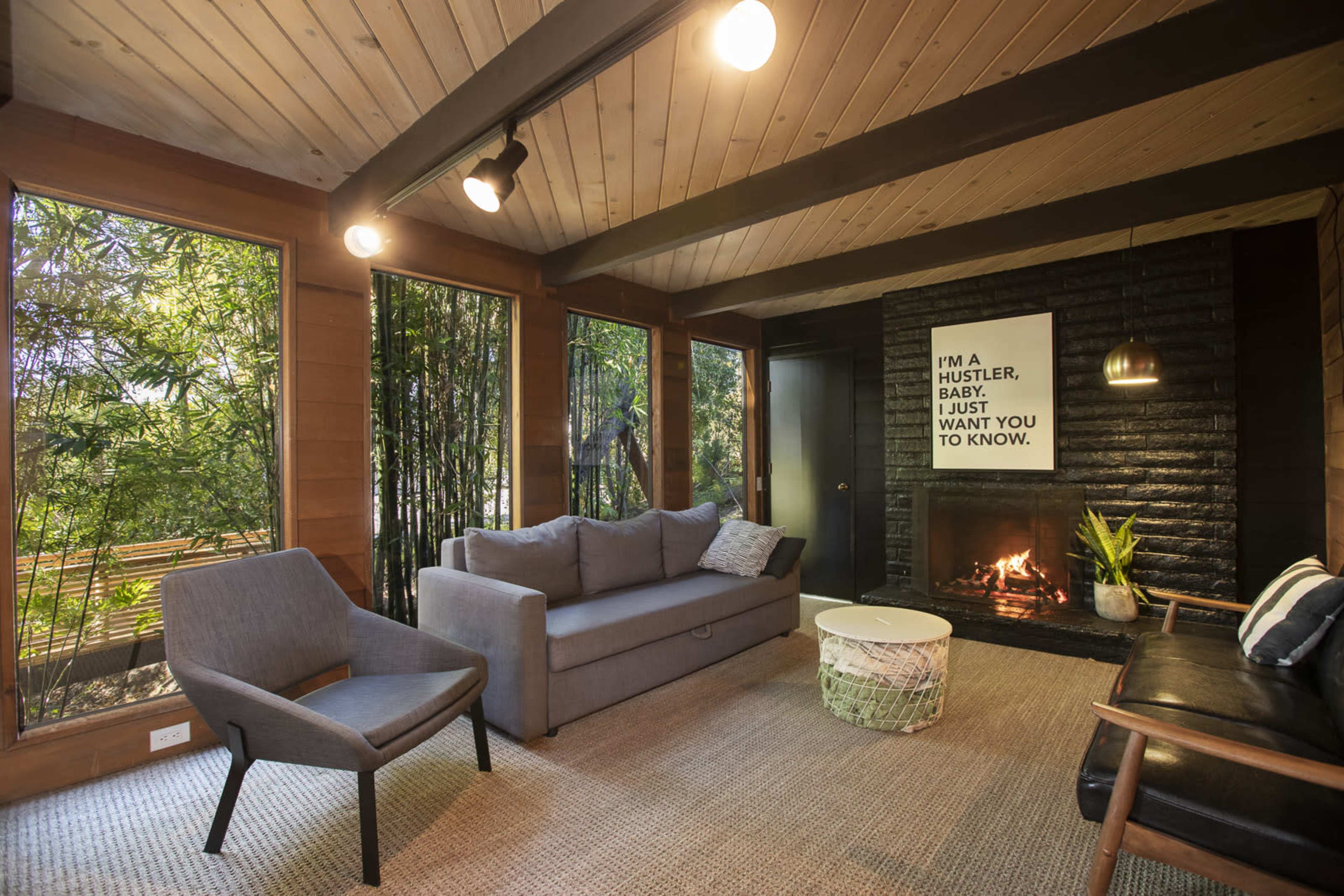 A living room features a sofa, armchair, and coffee table, with large windows that provide a view of greenery outside and a fireplace in the corner.