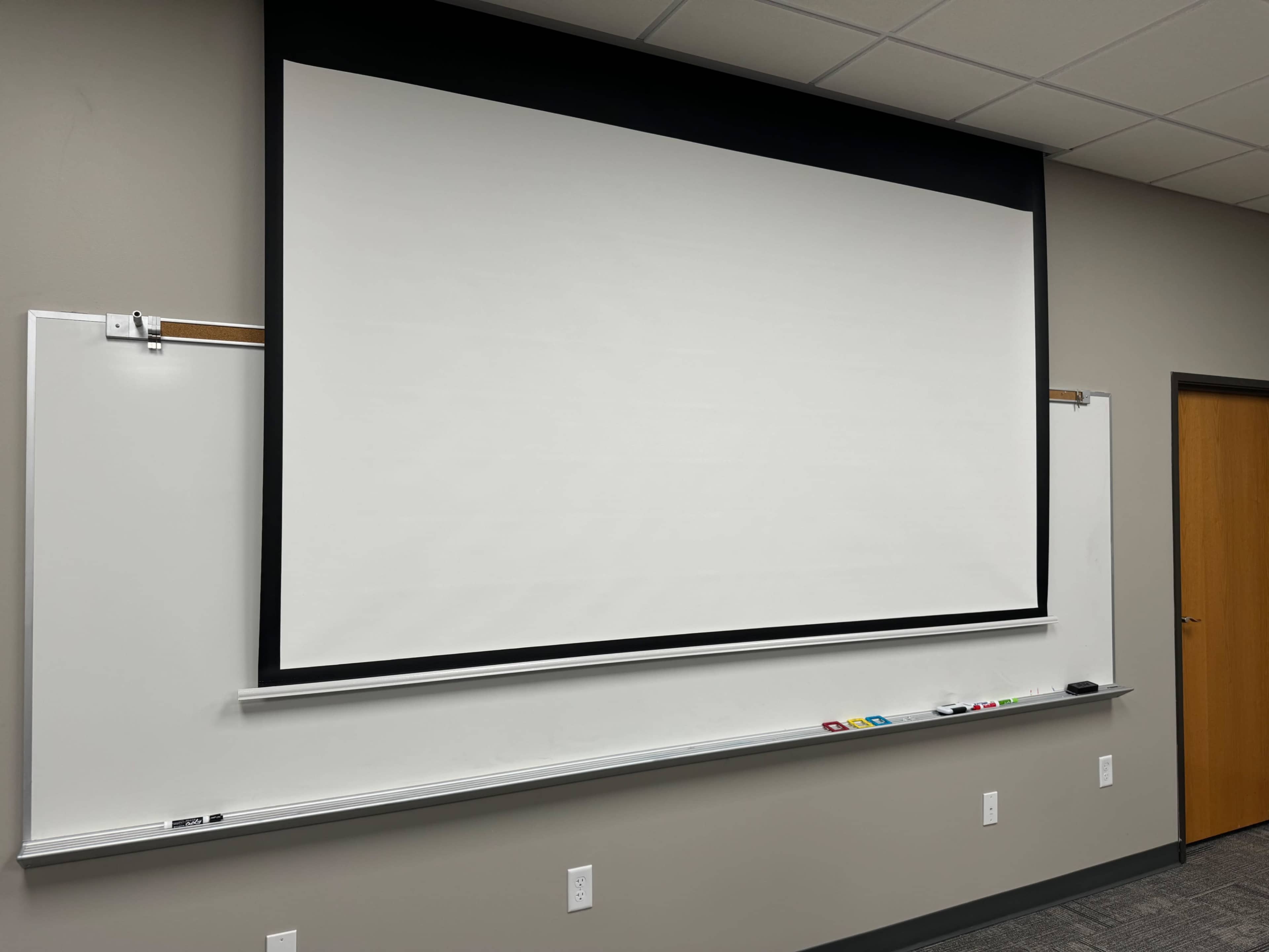 The image shows a classroom or presentation room with a blank projection screen mounted on a wall, accompanied by a whiteboard beneath it.