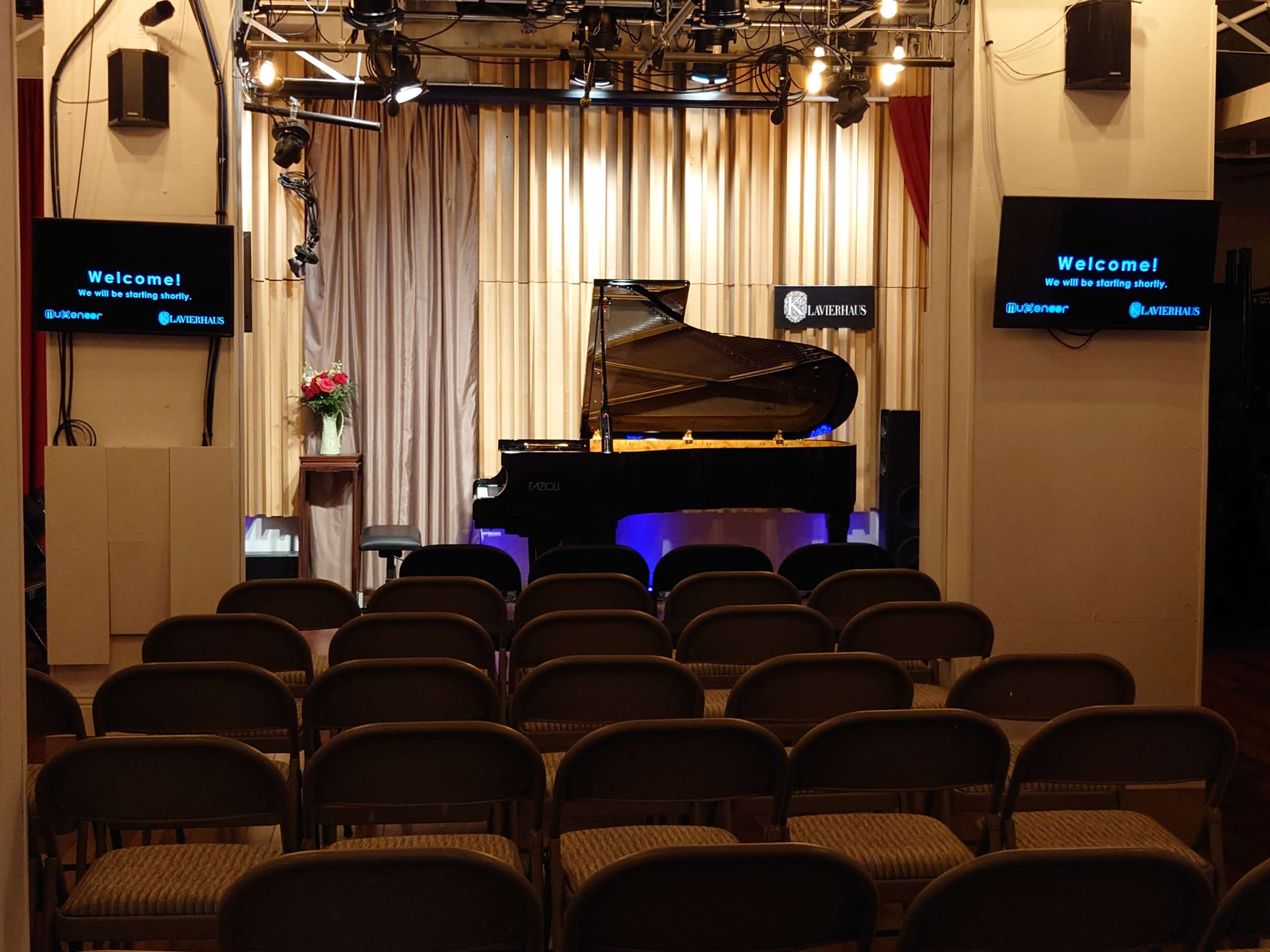 The image shows a small performance space with a grand piano on stage, surrounded by rows of chairs for an audience, and screens displaying a welcome message.