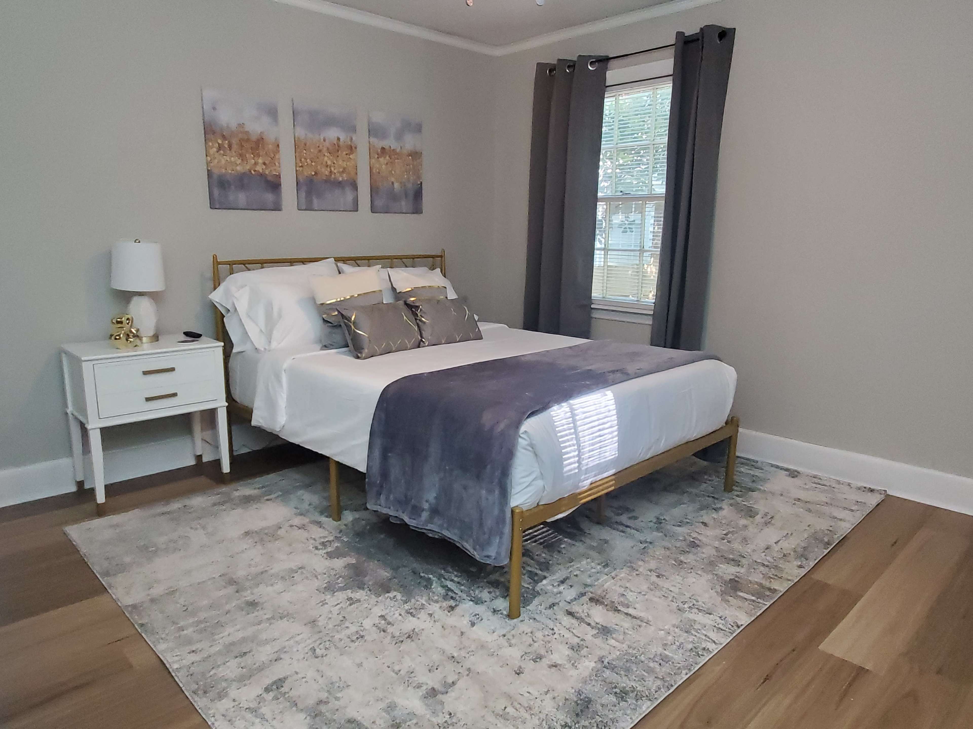 The image shows a neatly arranged bedroom featuring a bed with white linens, a gray throw, a nightstand, and three framed artworks on the wall.