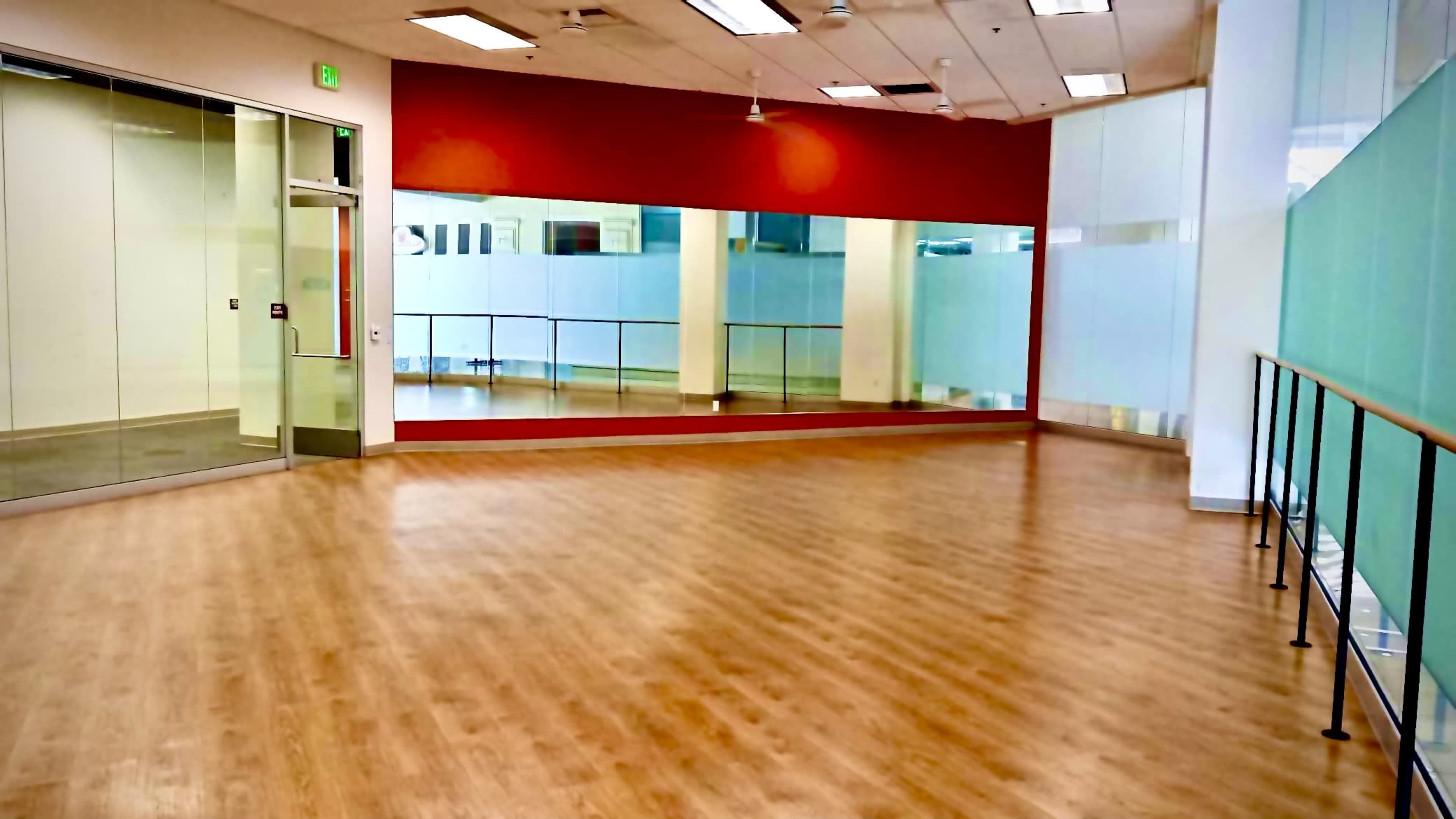 The image shows a spacious, empty room with wooden floors and large mirrors on one wall.