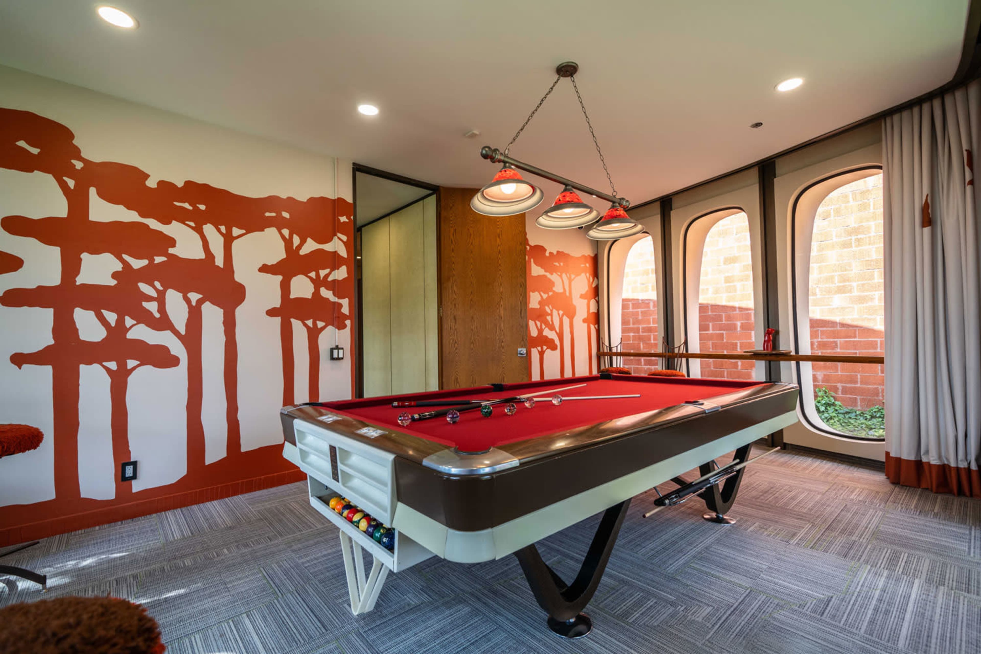 The image shows a game room featuring a red-felt pool table under a trio of pendant lights, with a wall adorned by a tree-themed mural.