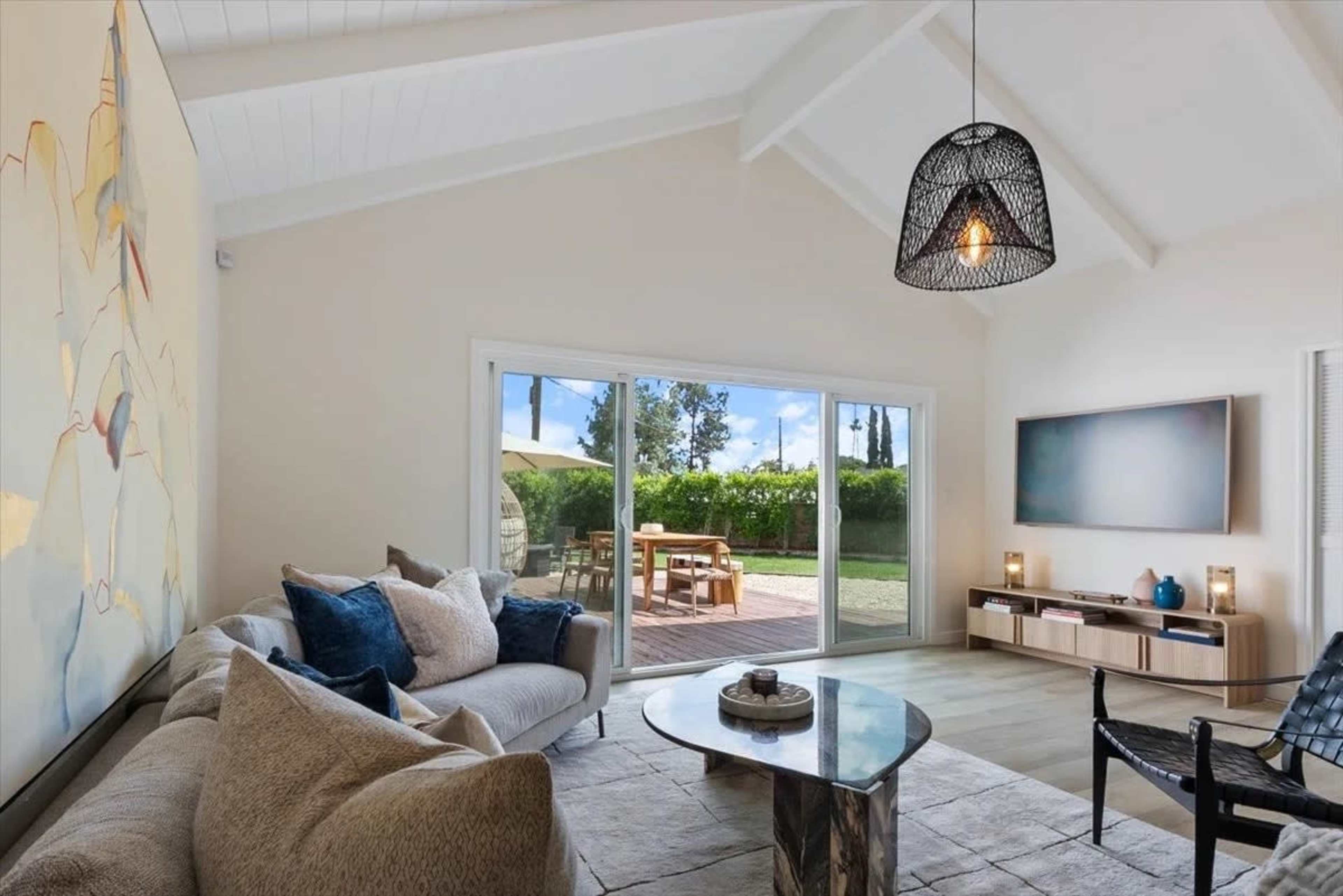 A modern living room features a large sectional sofa, a coffee table, and a wall-mounted television, with large sliding doors opening to a patio and view of greenery outside.