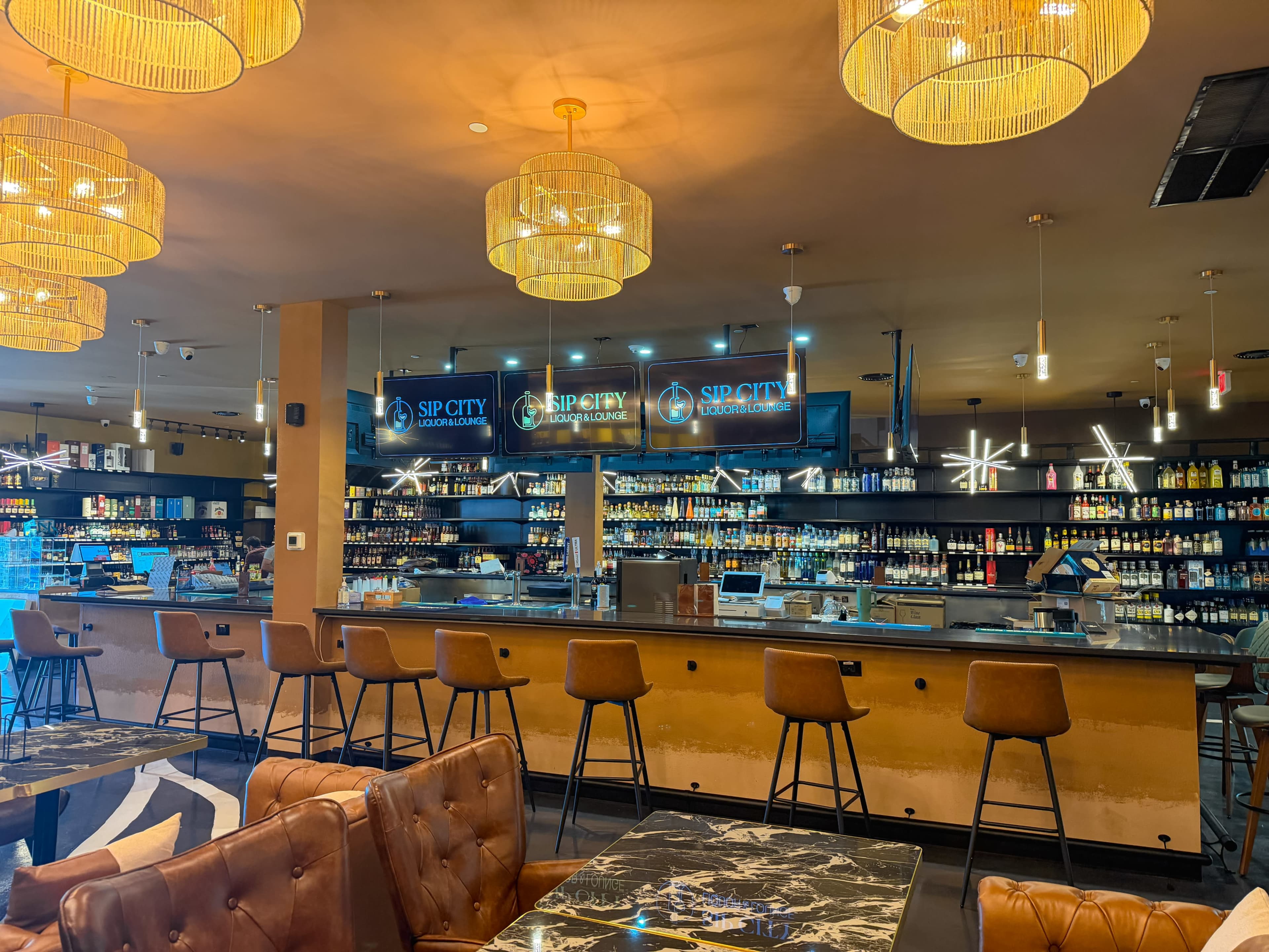 The image shows a modern bar area with a sleek countertop, high stools, and multiple illuminated shelves displaying various bottles of liquor.