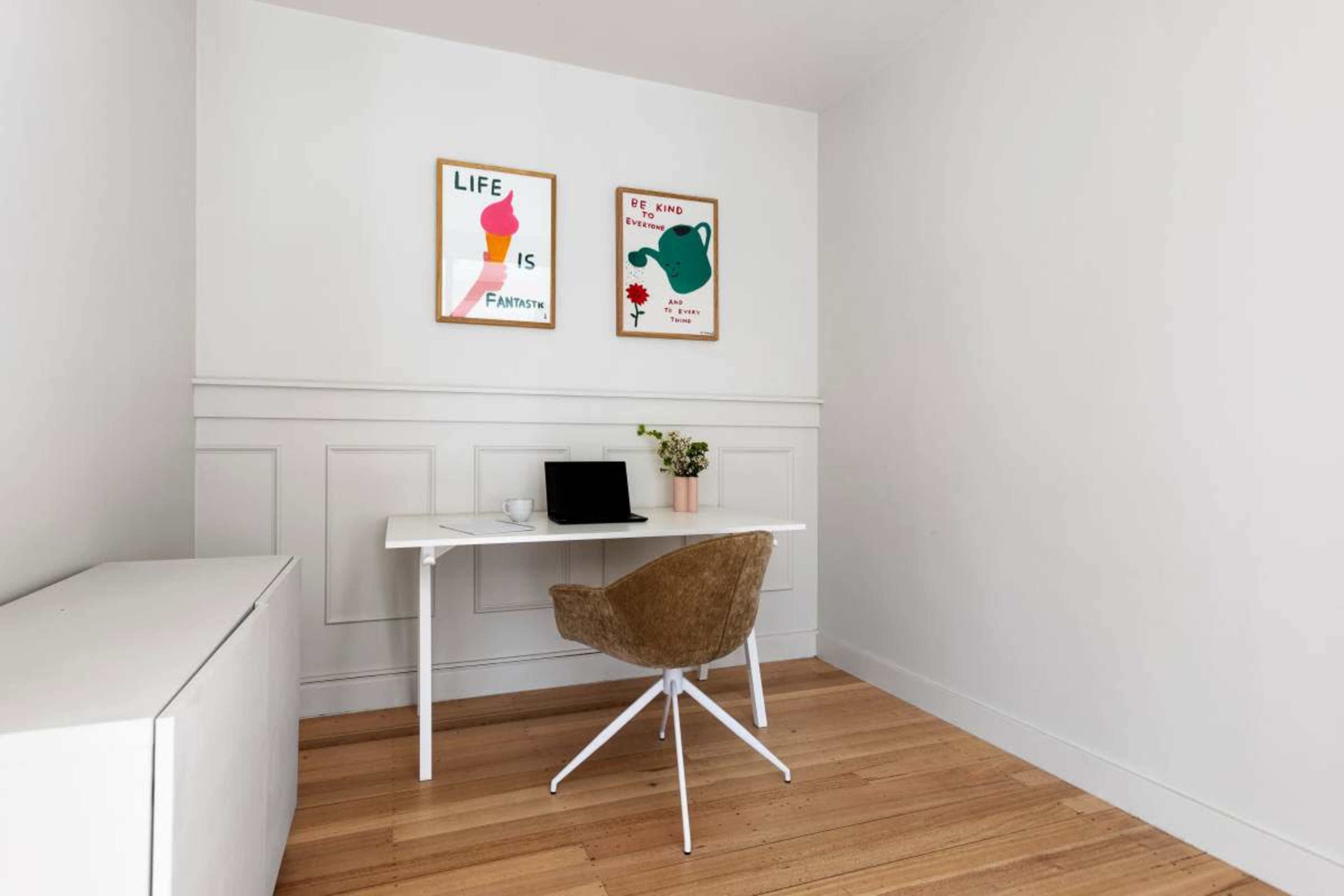 A minimalist home office features a white desk with a laptop, a brown chair, and two framed posters on the wall, alongside a small potted plant.