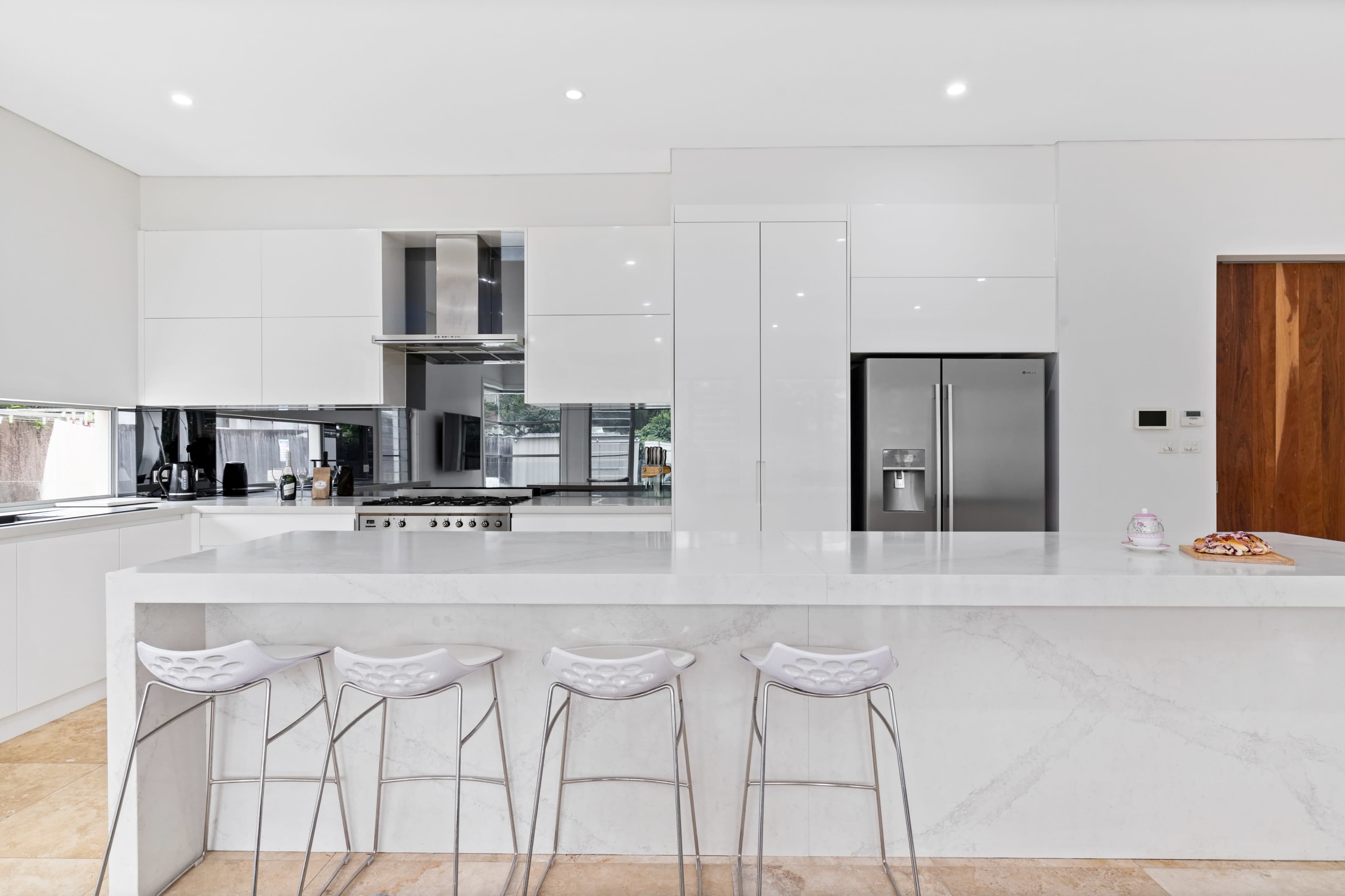 The image shows a modern kitchen featuring a large island with four bar stools, sleek white cabinets, stainless steel appliances, and a natural stone countertop.