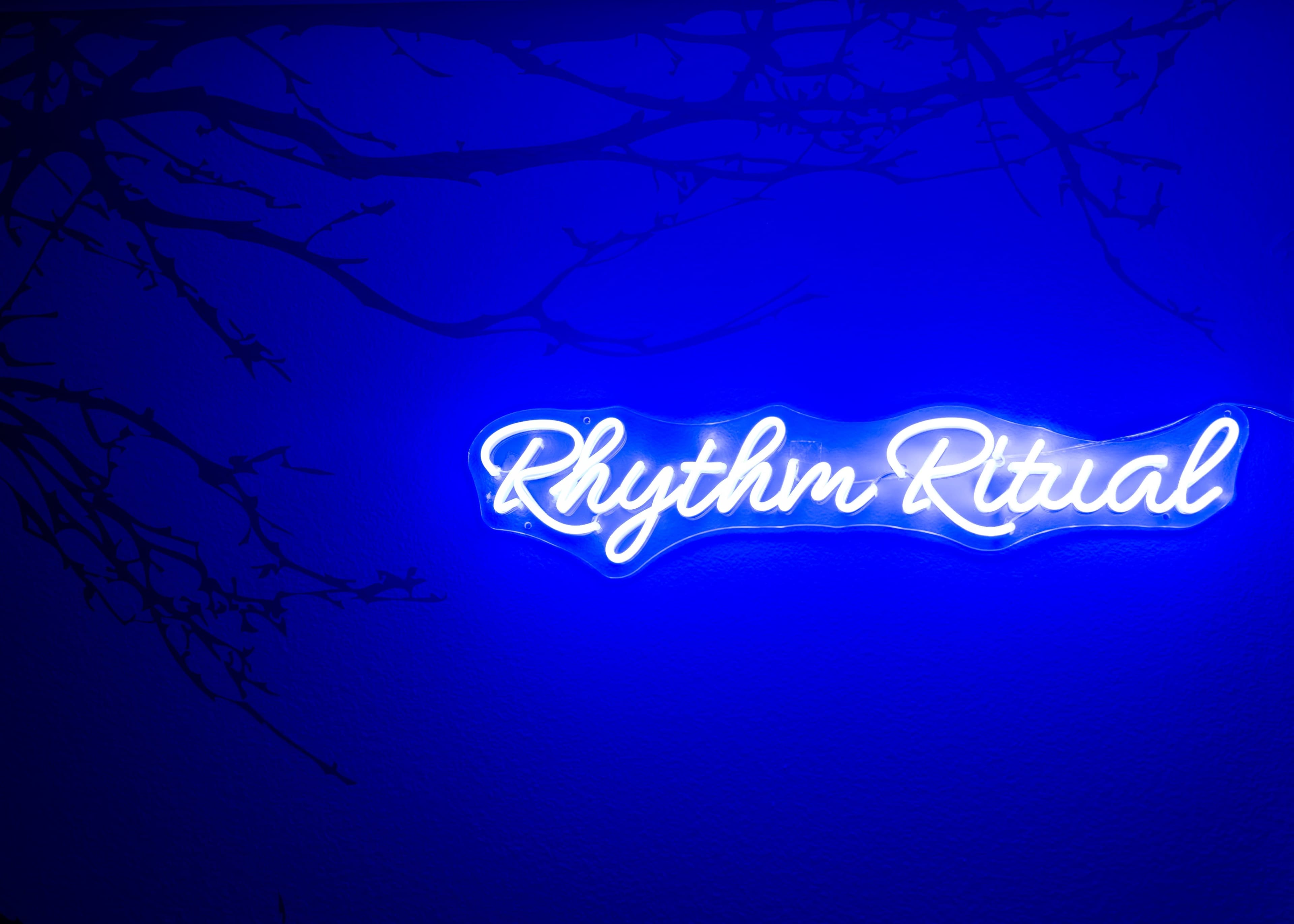 A glowing blue neon sign displaying the words "Rhythm Ritual" is mounted on a wall, with a dark, shadowy outline of branches in the background.