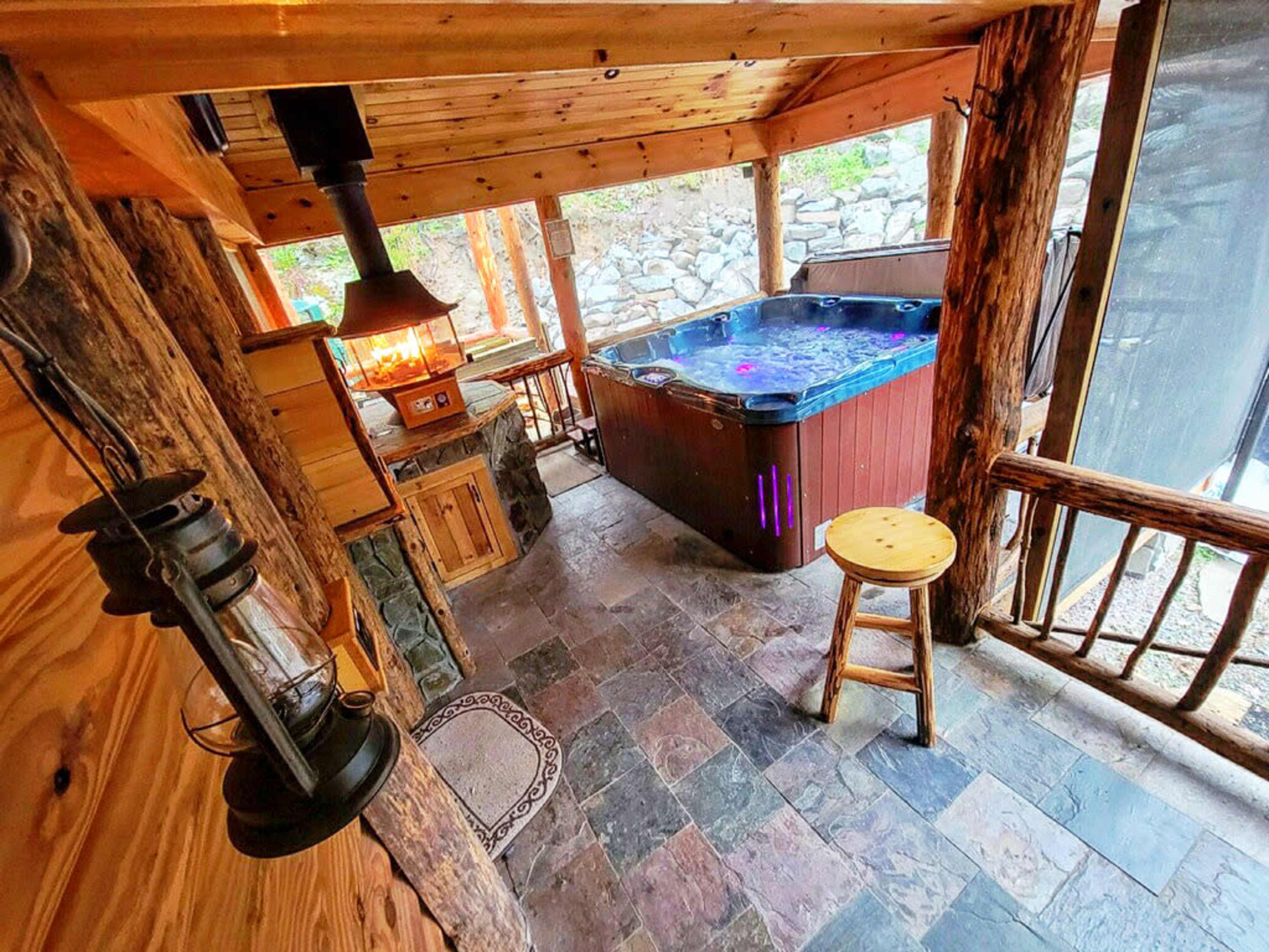 The image shows a wooden cabin interior featuring a hot tub, a stone fireplace, and rustic furnishings.