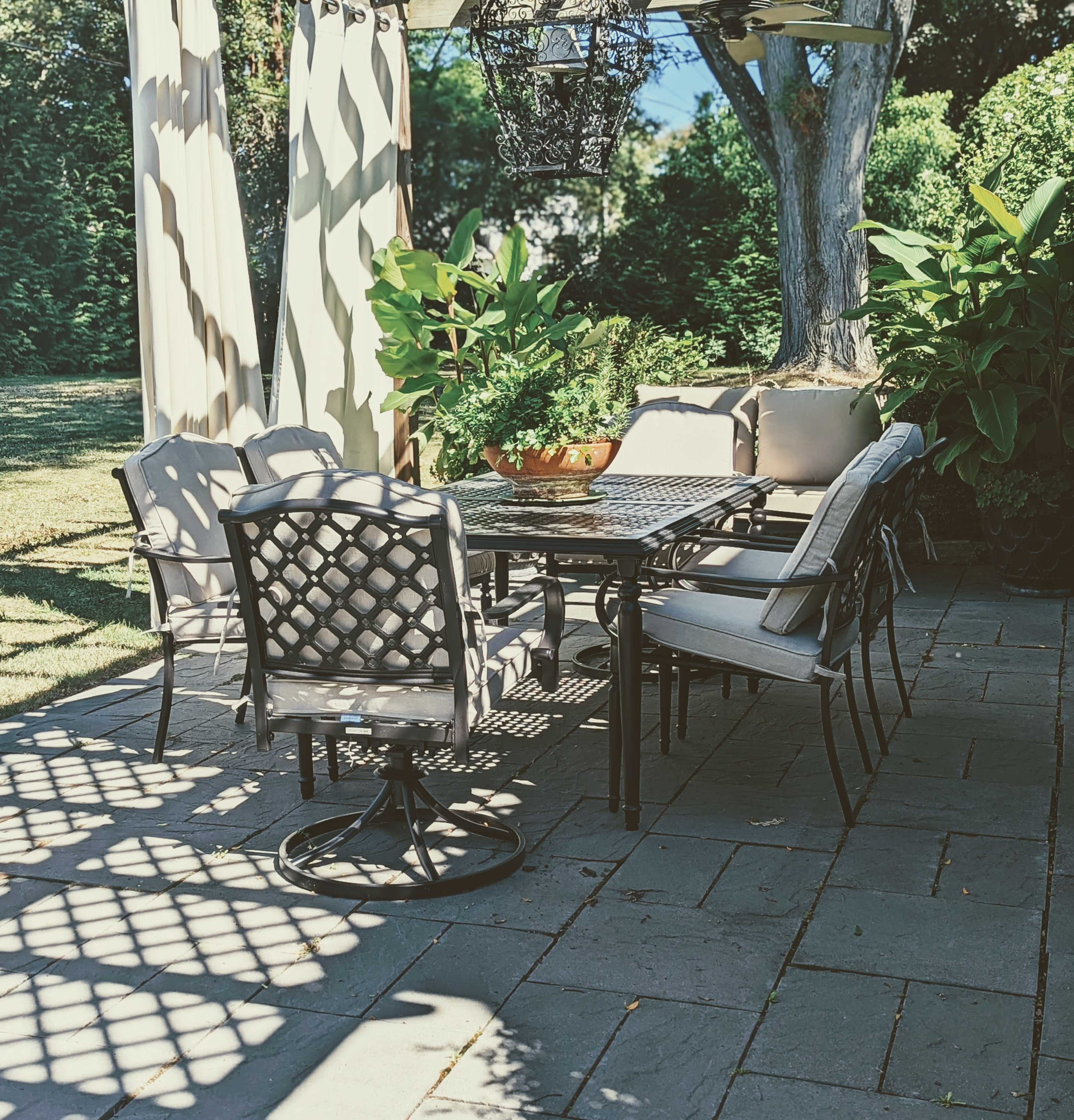 A patio seating area features a metal dining table surrounded by six chairs, shaded by a pergola and illuminated by hanging lanterns, with greenery in the background.