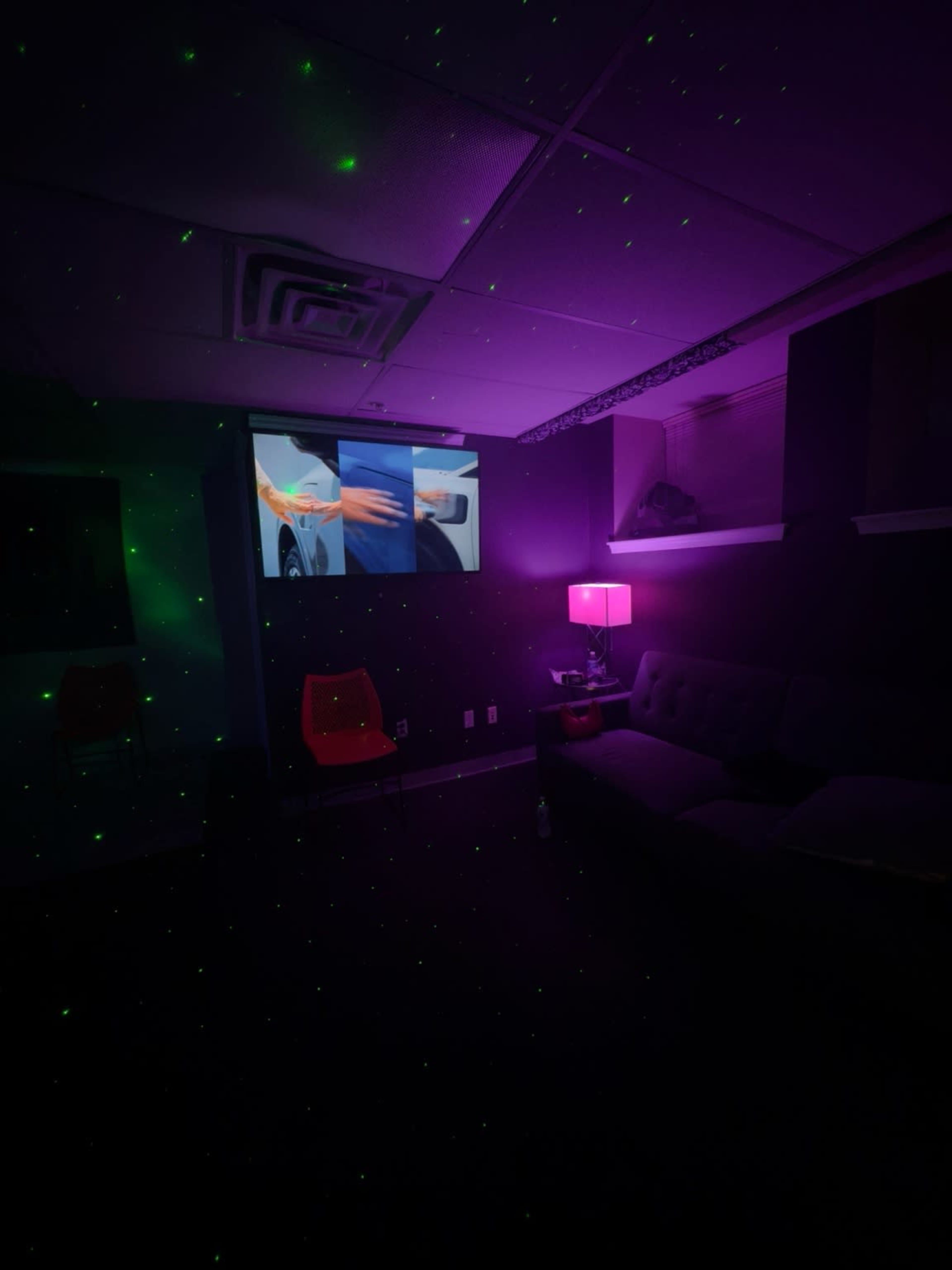A dimly lit room features a couch and a purple lamp, with two screens displaying videos on a wall, surrounded by green laser dots.