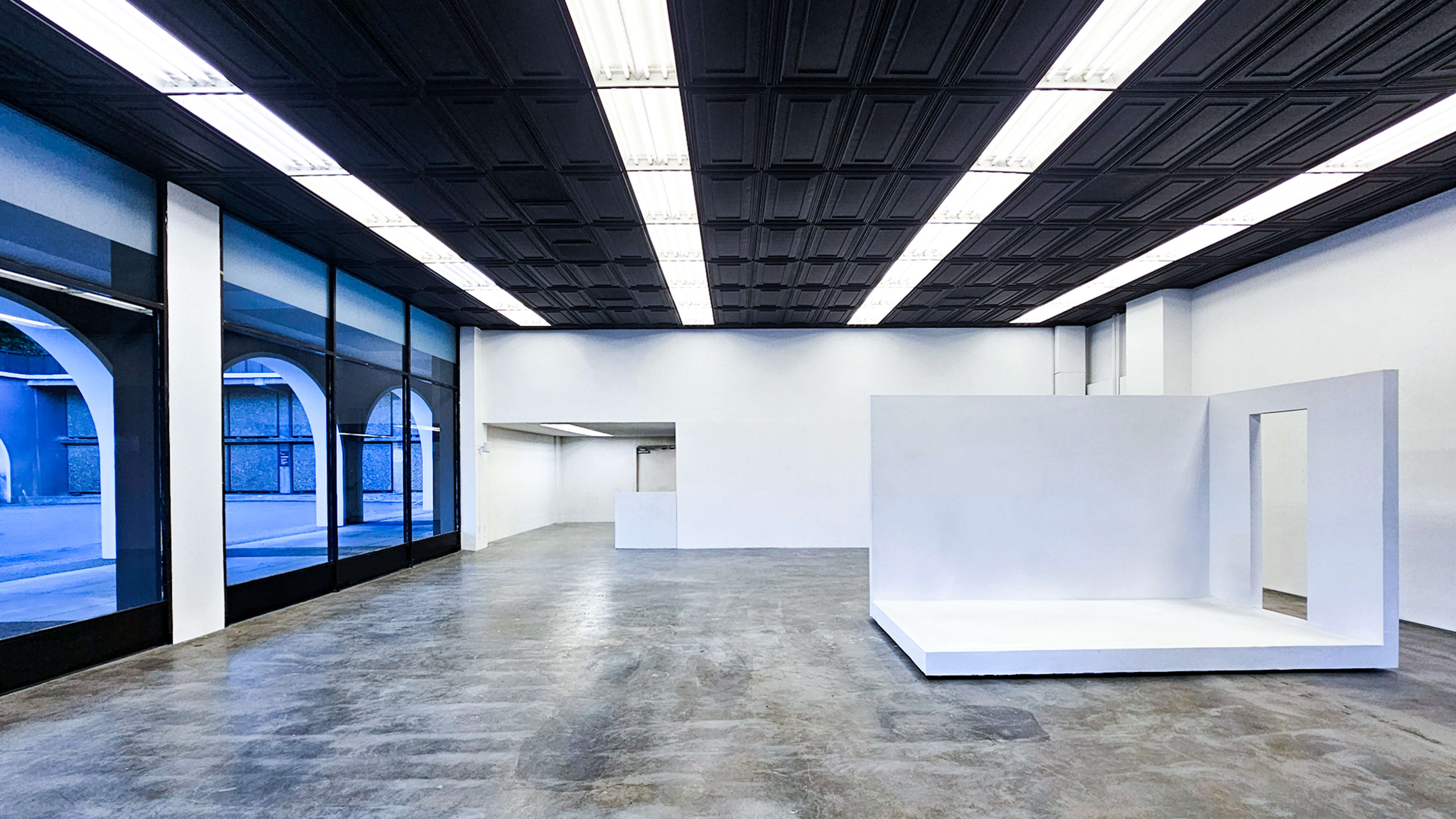 The image shows an empty gallery space with large windows, a polished concrete floor, and a white, minimalistic structure in the center.