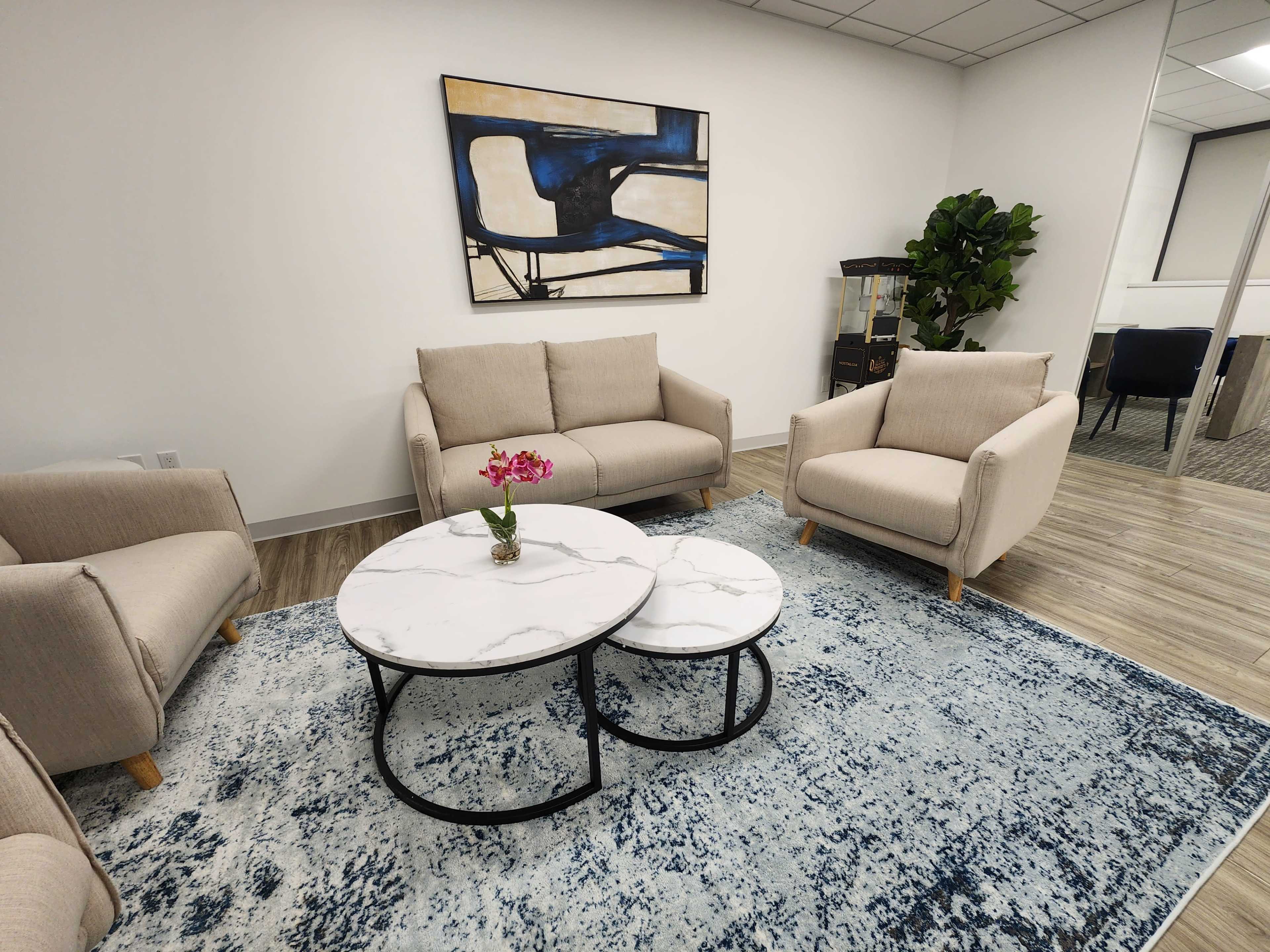 A modern office seating area with two beige sofas and a round marble coffee table on a blue rug, accompanied by a small plant and a wall painting.