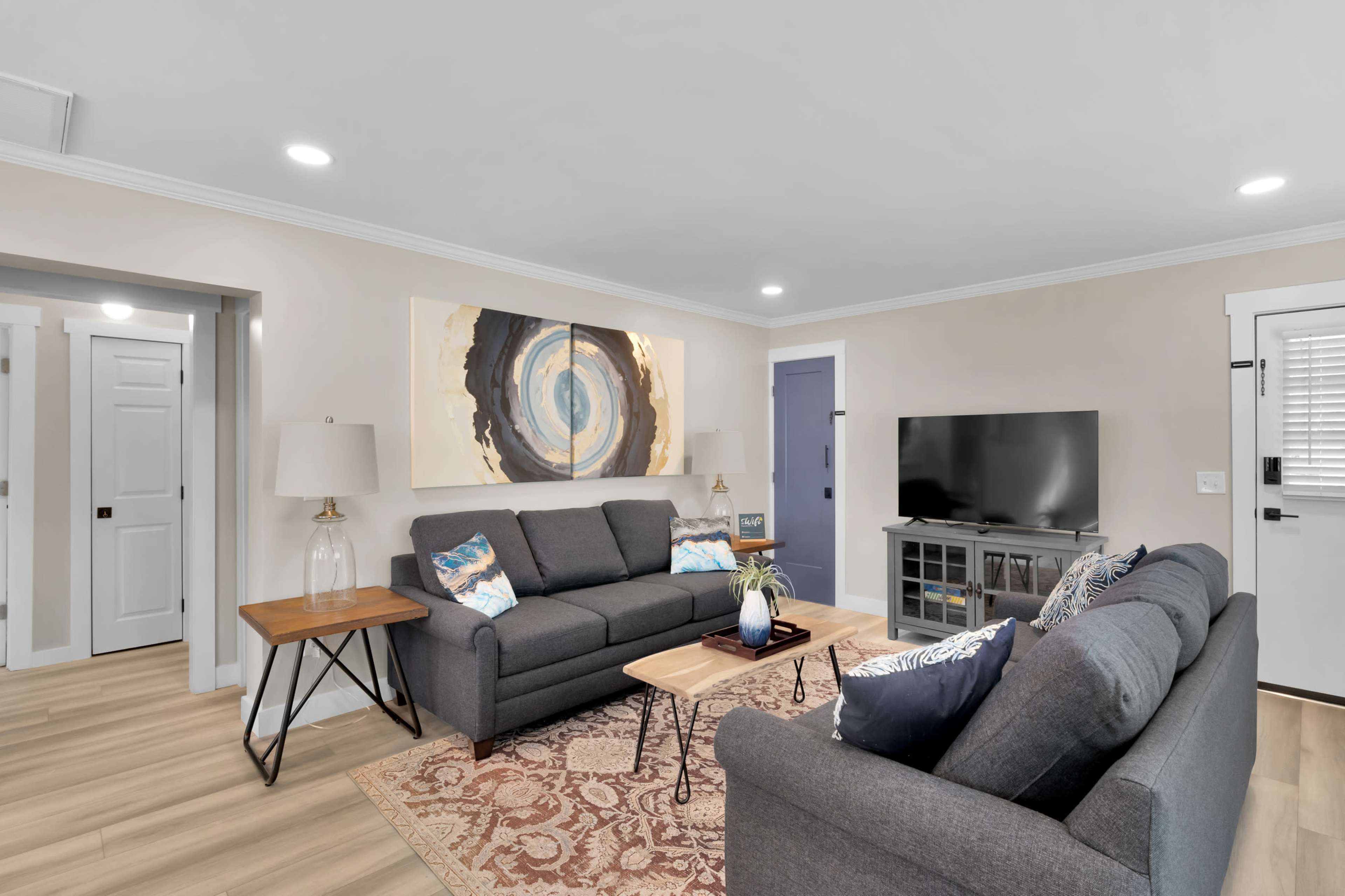 A living room features two gray sofas, a wooden coffee table, a television on a stand, and artwork on the wall.