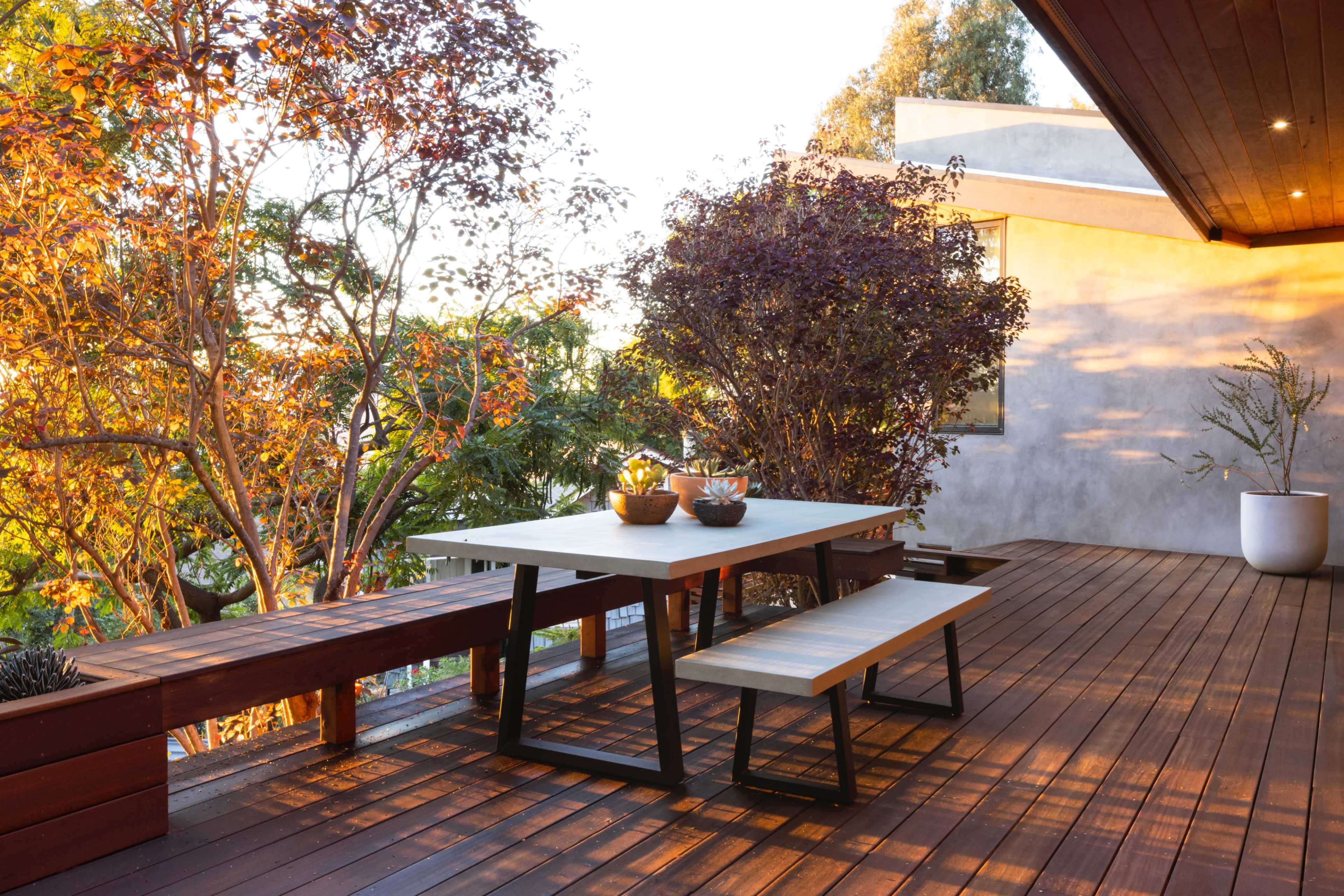 A modern wooden deck features a table and benches beside vibrant trees and plants.
