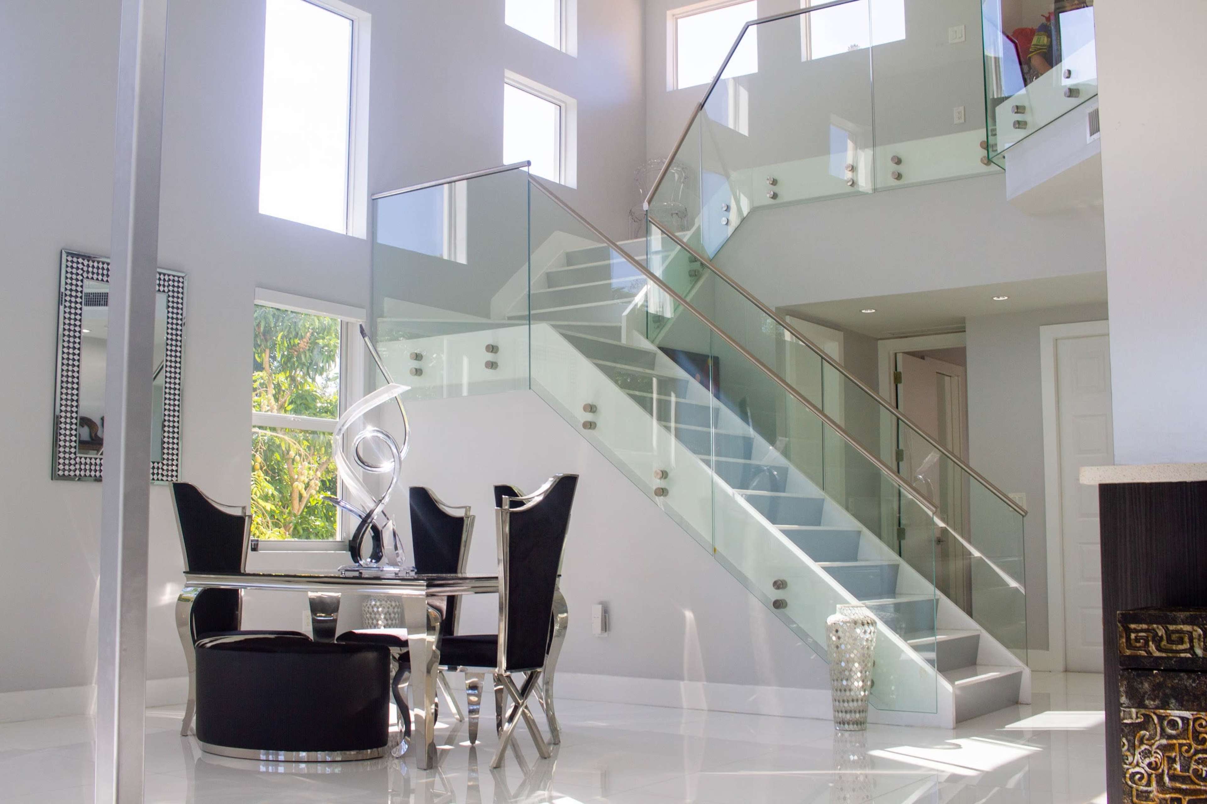 A modern indoor space featuring a glass staircase, a dining table with black chairs, and large windows allowing natural light.