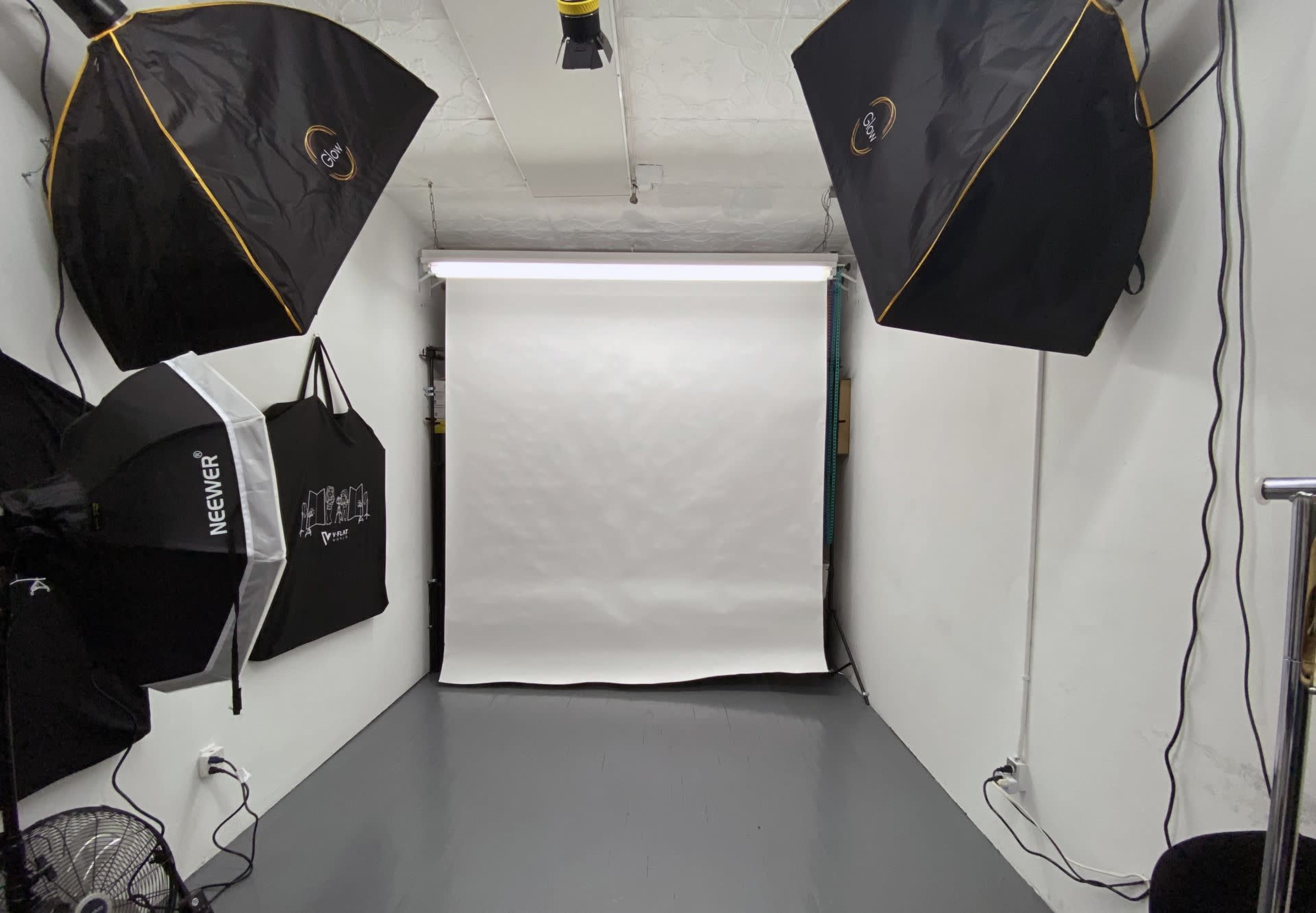The image shows a photography studio with two softbox lights, a backdrop stand with a white backdrop, and a floor fan.