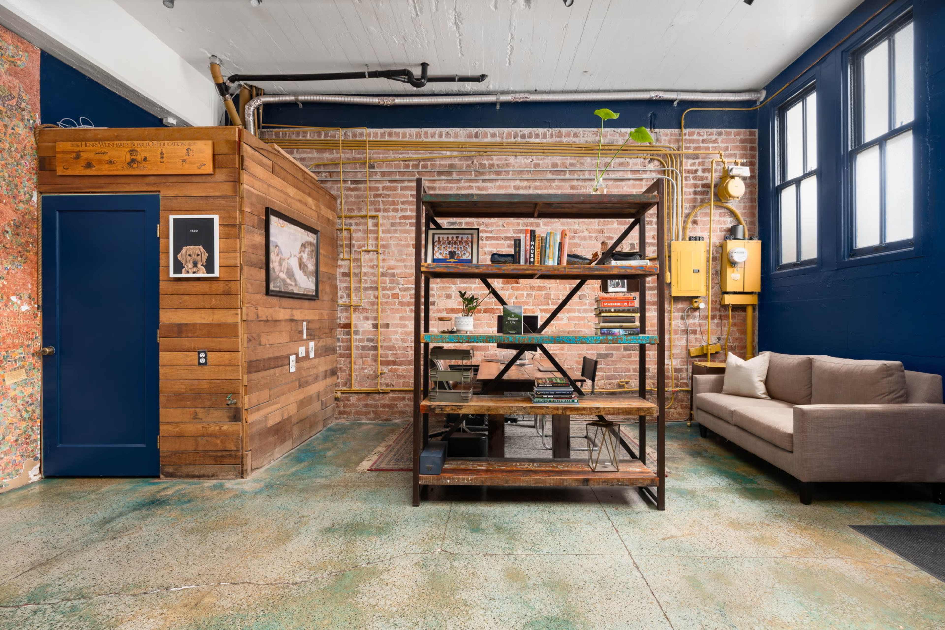 The image shows a modern industrial-style room with exposed brick walls, a wooden feature wall, a metal shelving unit filled with books, and a gray sofa.