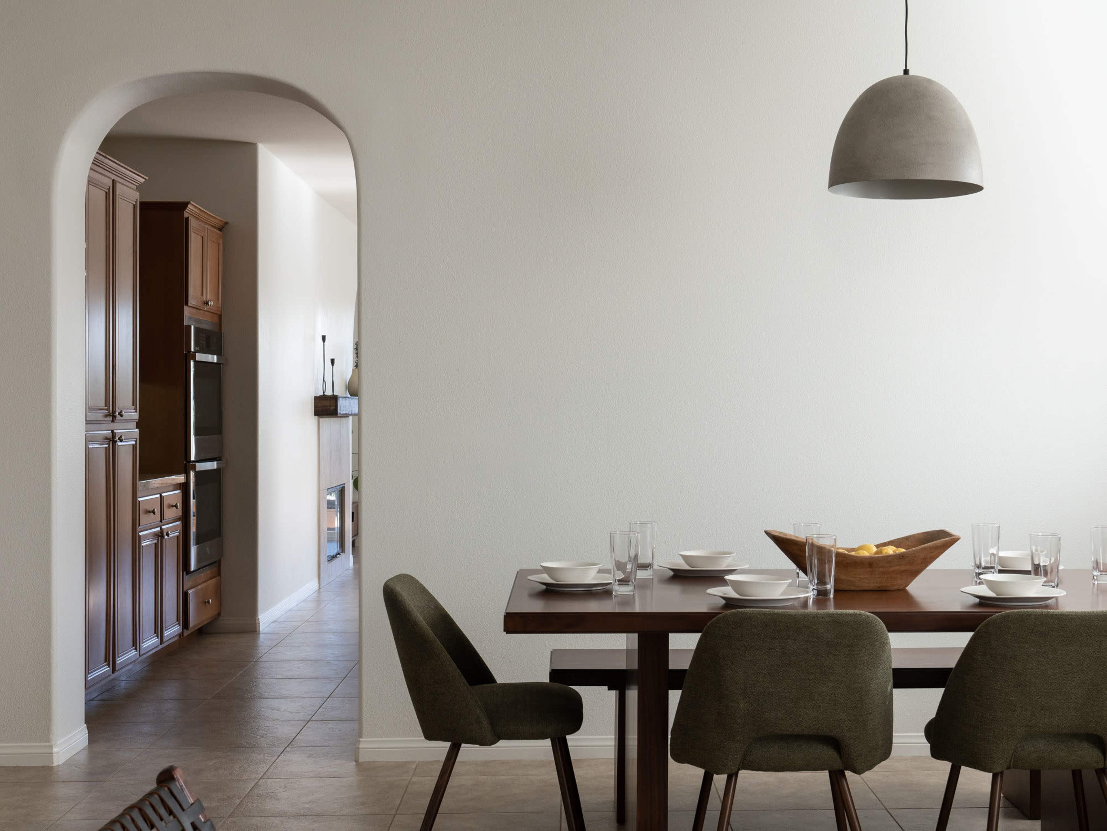 A dining area features a wooden table set with plates and glasses, with an archway leading to a kitchen in the background.