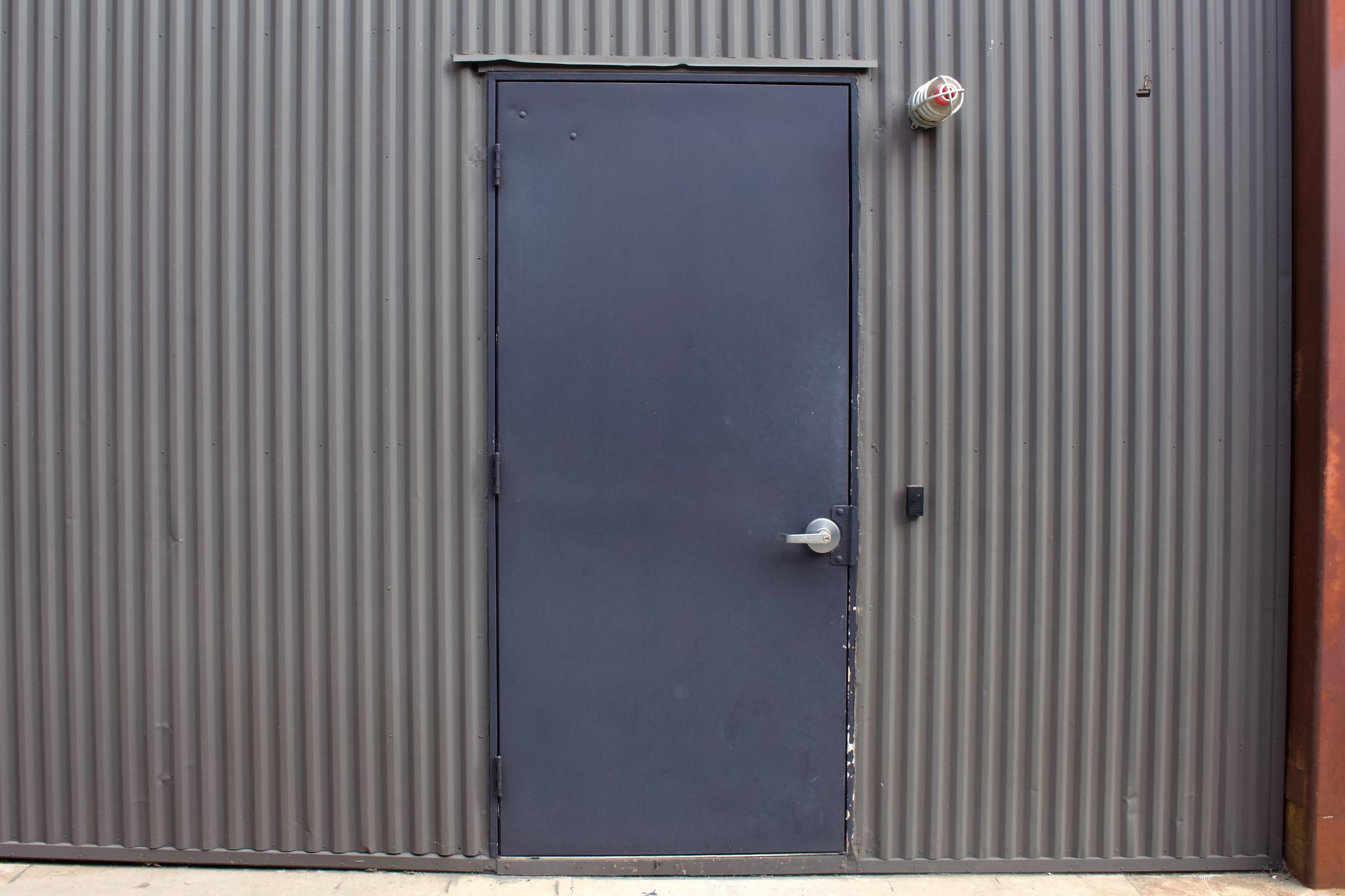 The image shows a plain gray metal door set against a corrugated metal wall.