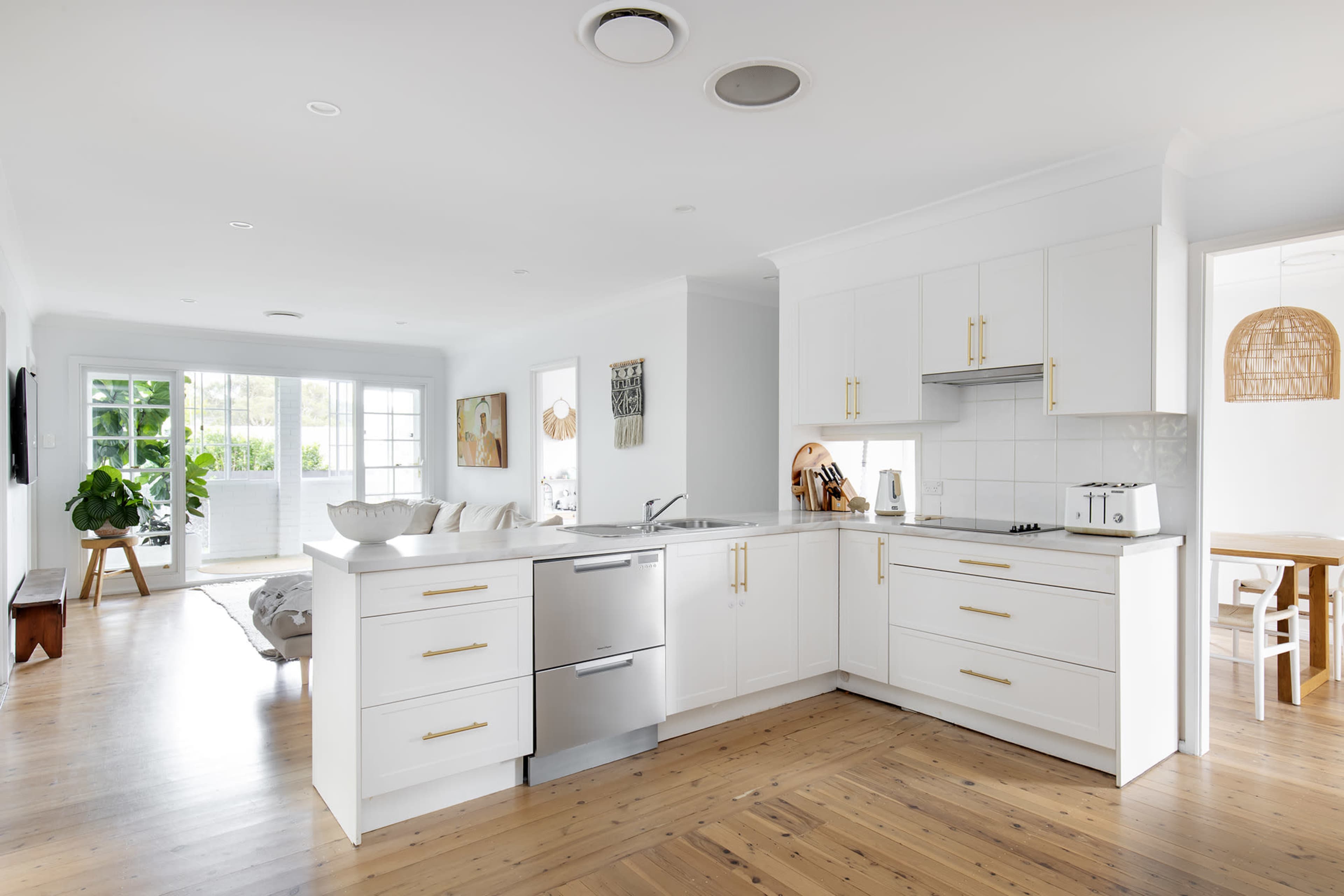 A modern kitchen features white cabinetry, a stainless steel dishwasher, and hardwood flooring, with an open layout connecting to a living area.