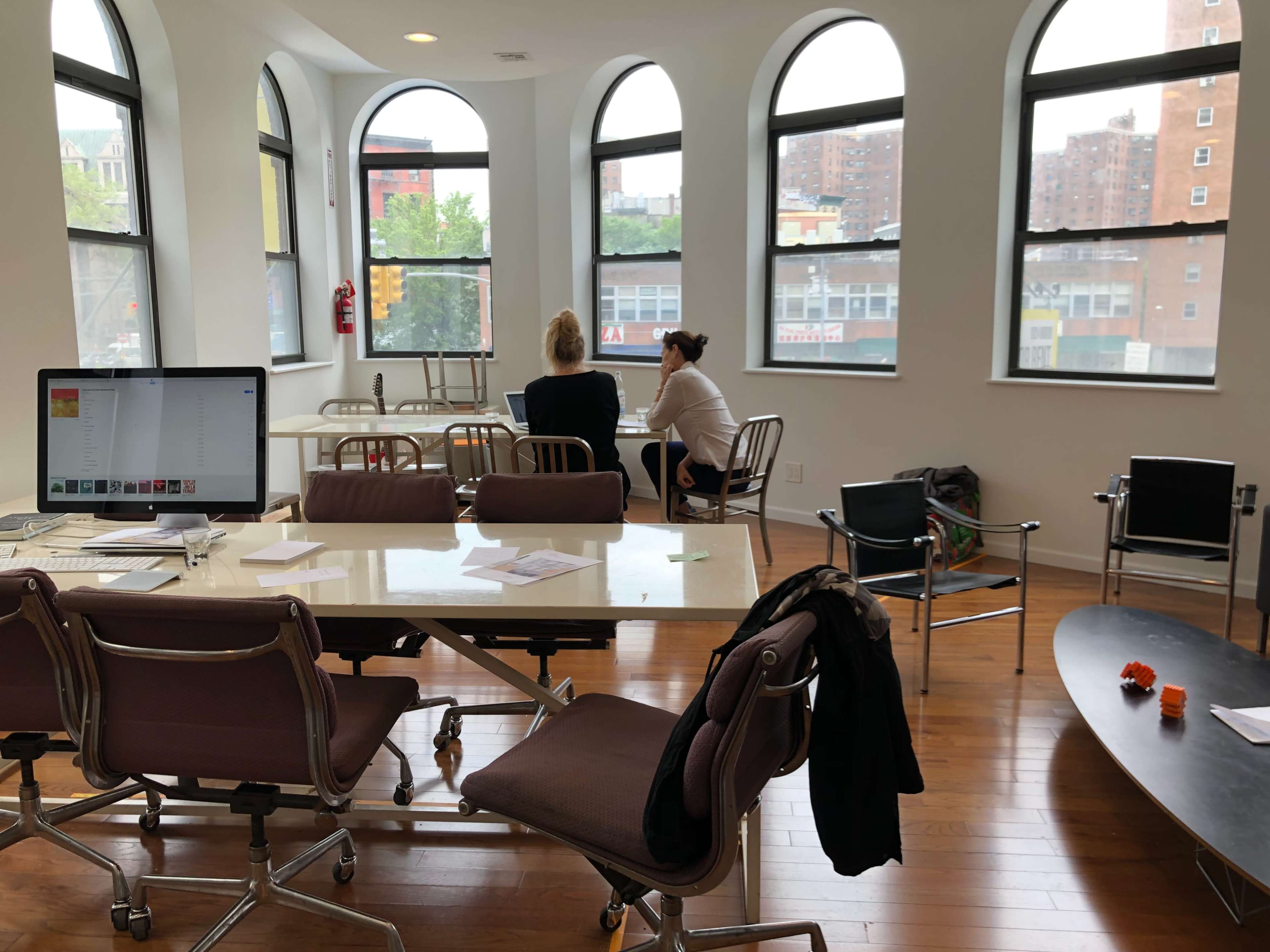 The image shows a modern office space with large arched windows, two people seated at a table, and a computer on a desk in the foreground.