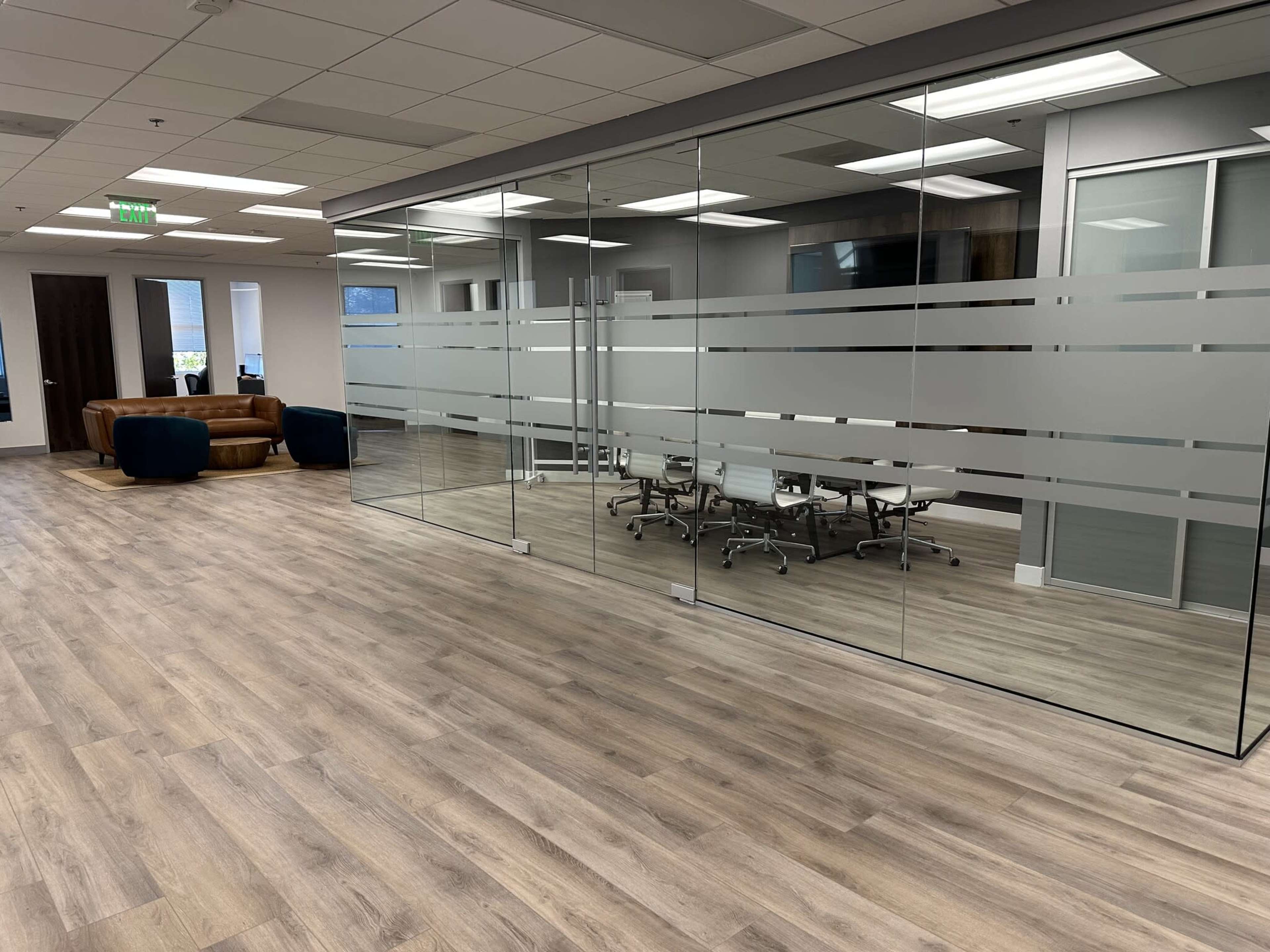 The image shows a modern office space with a glass-walled conference room, wooden flooring, and a seating area featuring a brown sofa and blue chairs.