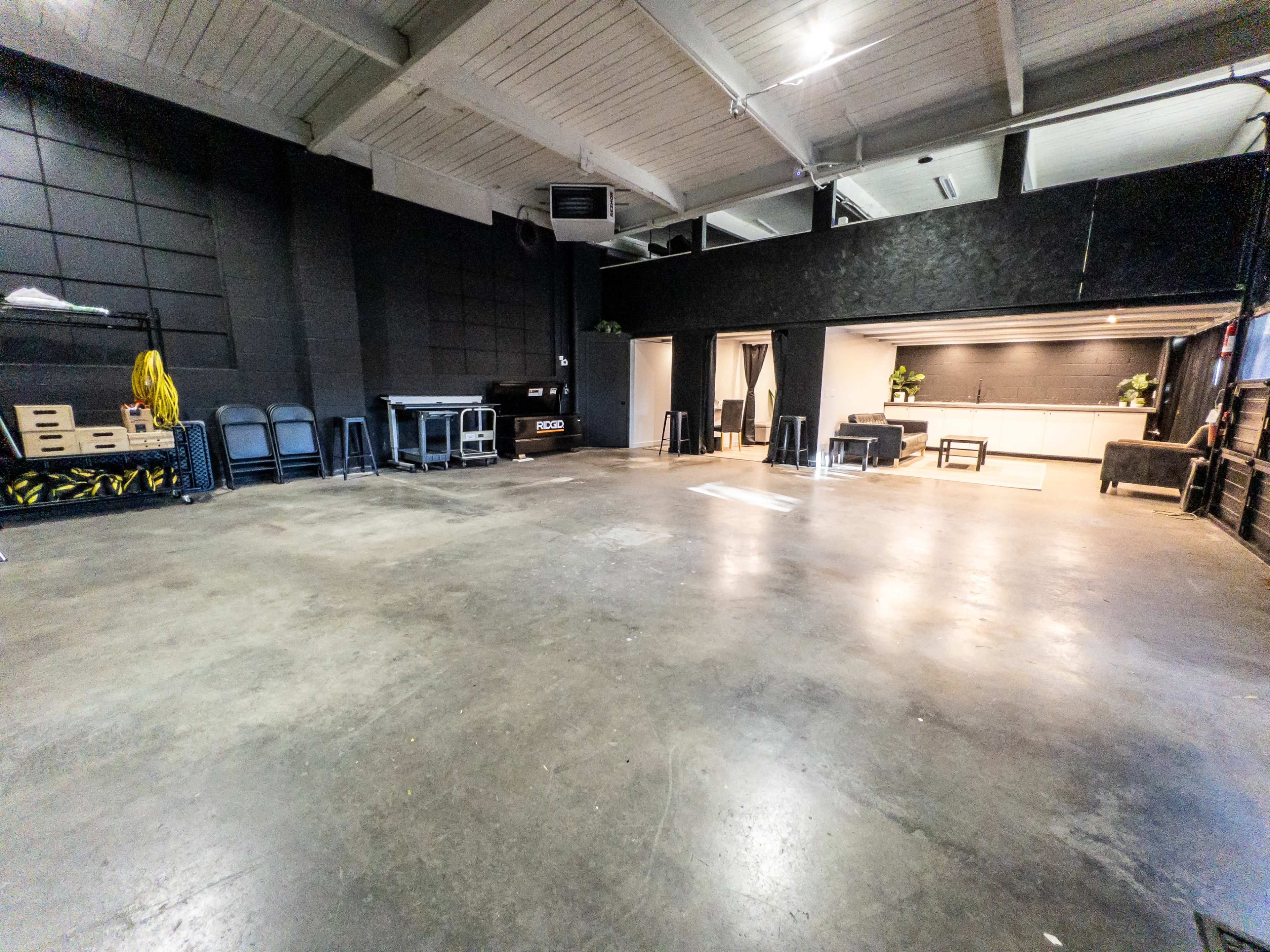 A spacious, empty industrial room with polished concrete floors, black walls, and minimalist furnishings in one corner.