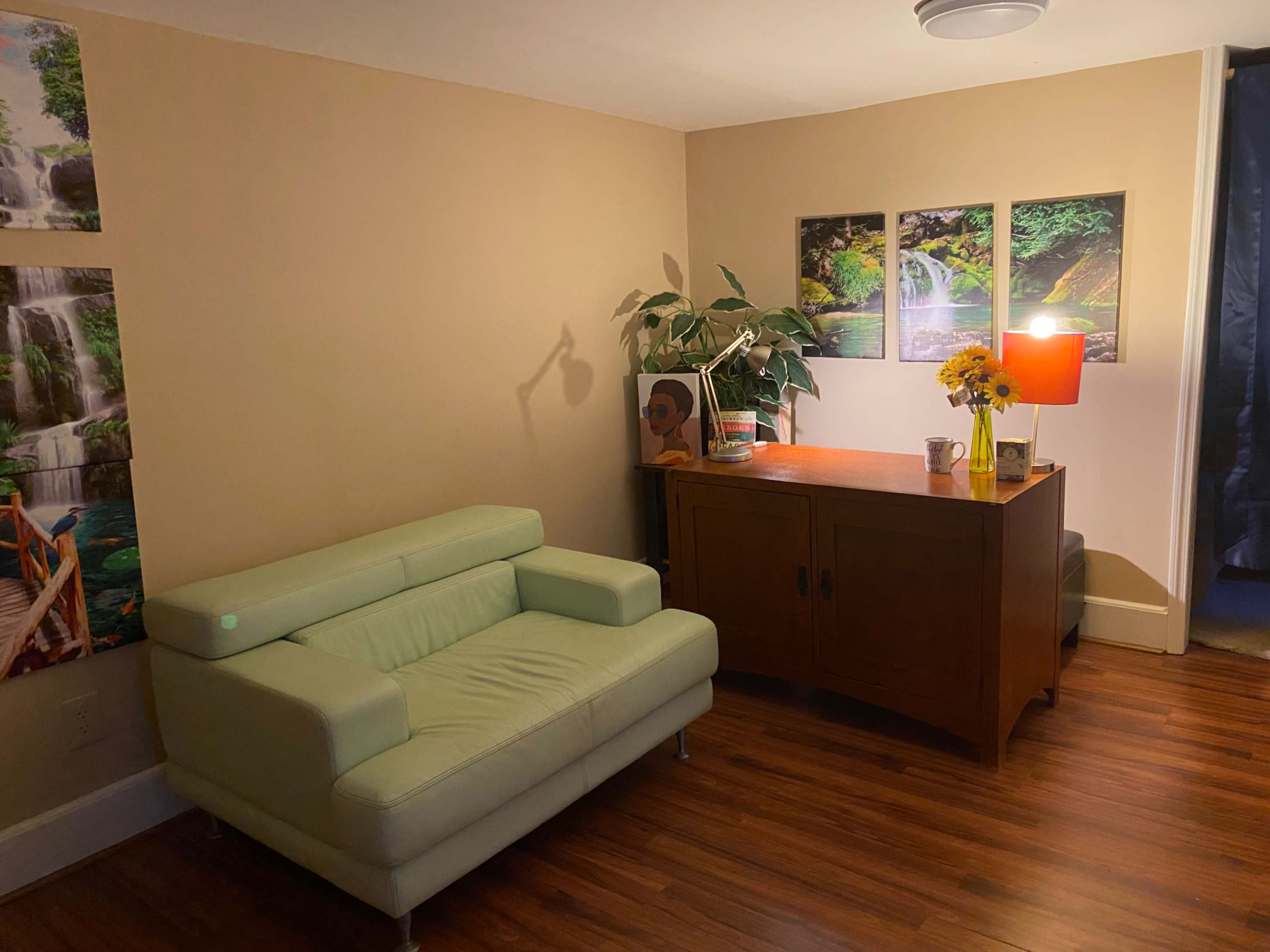 A small living room with a light green sofa, a wooden side table, a lamp, a vase of flowers, and paintings of waterfalls on the wall.