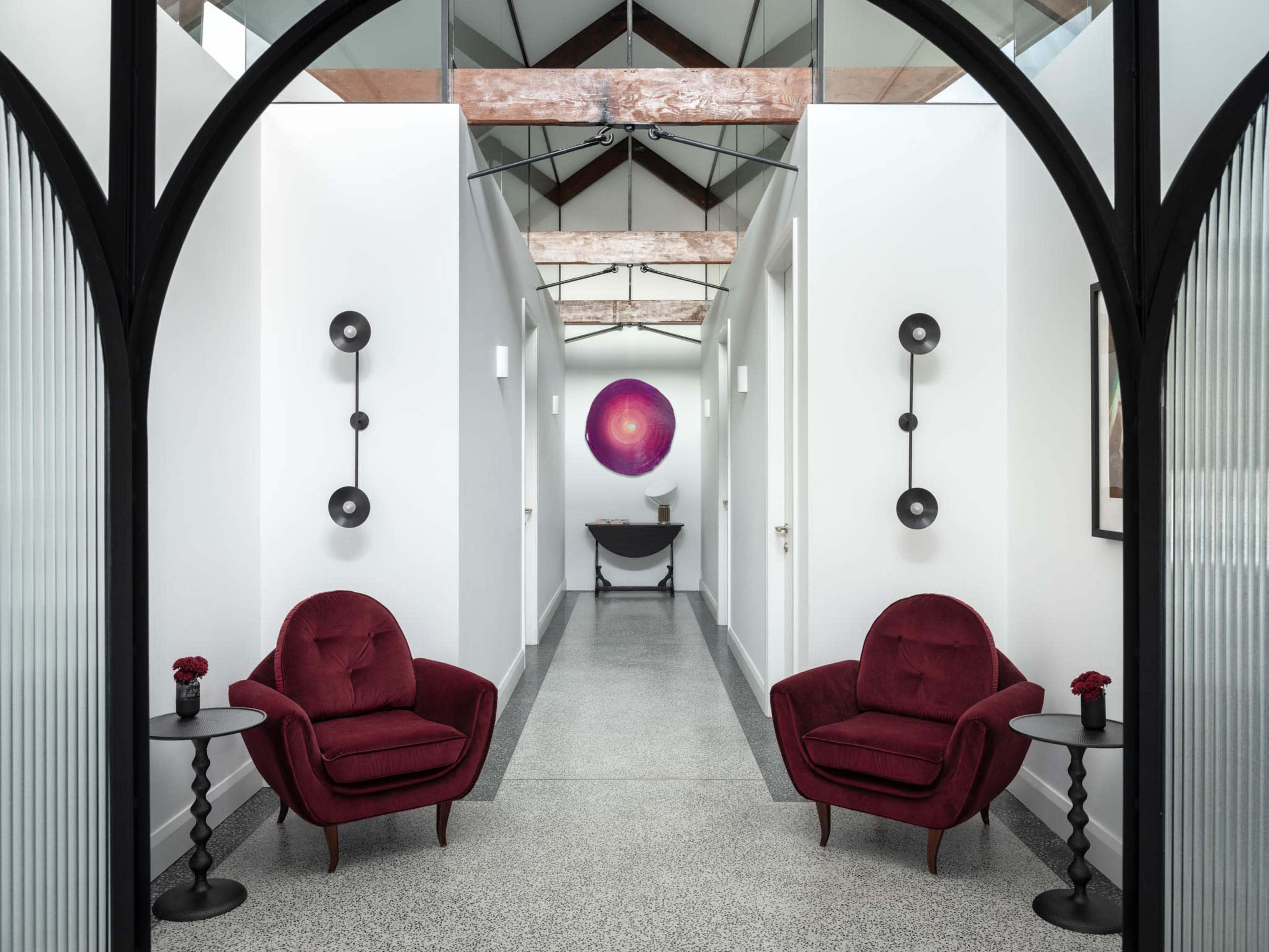 The image shows a modern hallway featuring two red velvet chairs and a circular artwork at the end.