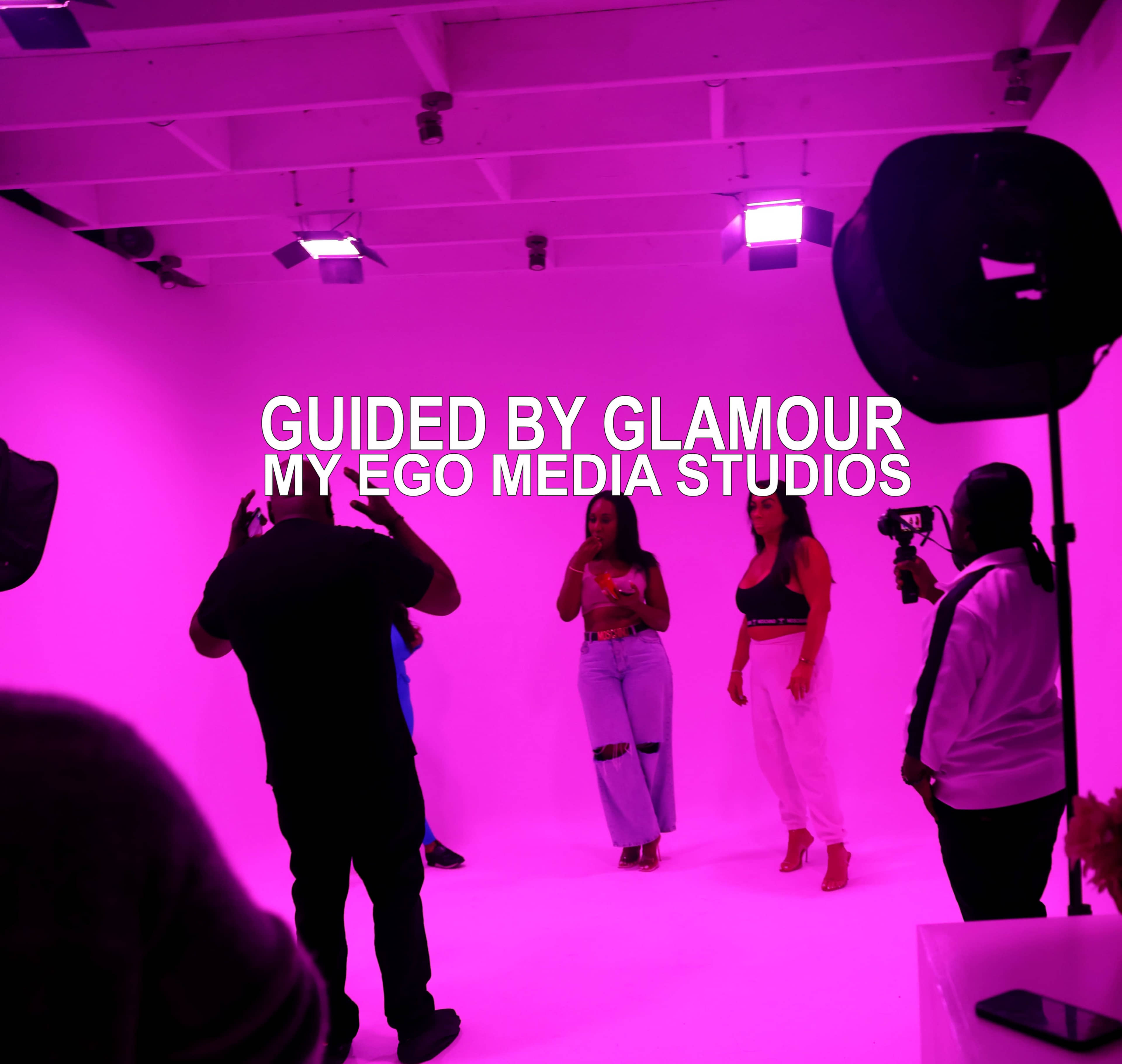 The image shows a group of people in a brightly lit studio with a pink backdrop, participating in a photoshoot or video production, with a banner reading "GUIDED BY GLAMOUR MY EGO MEDIA STUDIOS."