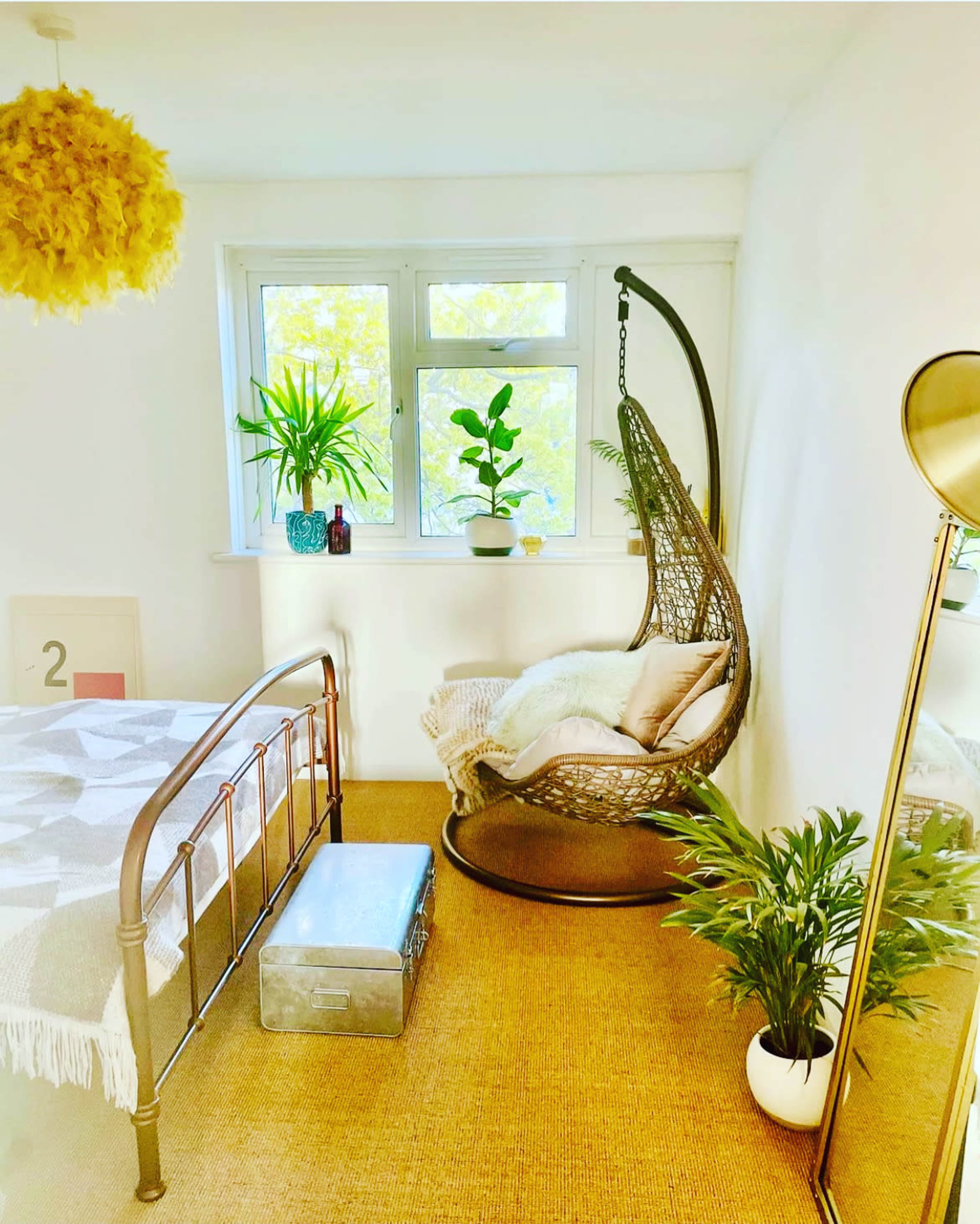 The image features a cozy bedroom with a metal bed, a hanging rattan chair, potted plants, and a decorative lamp.