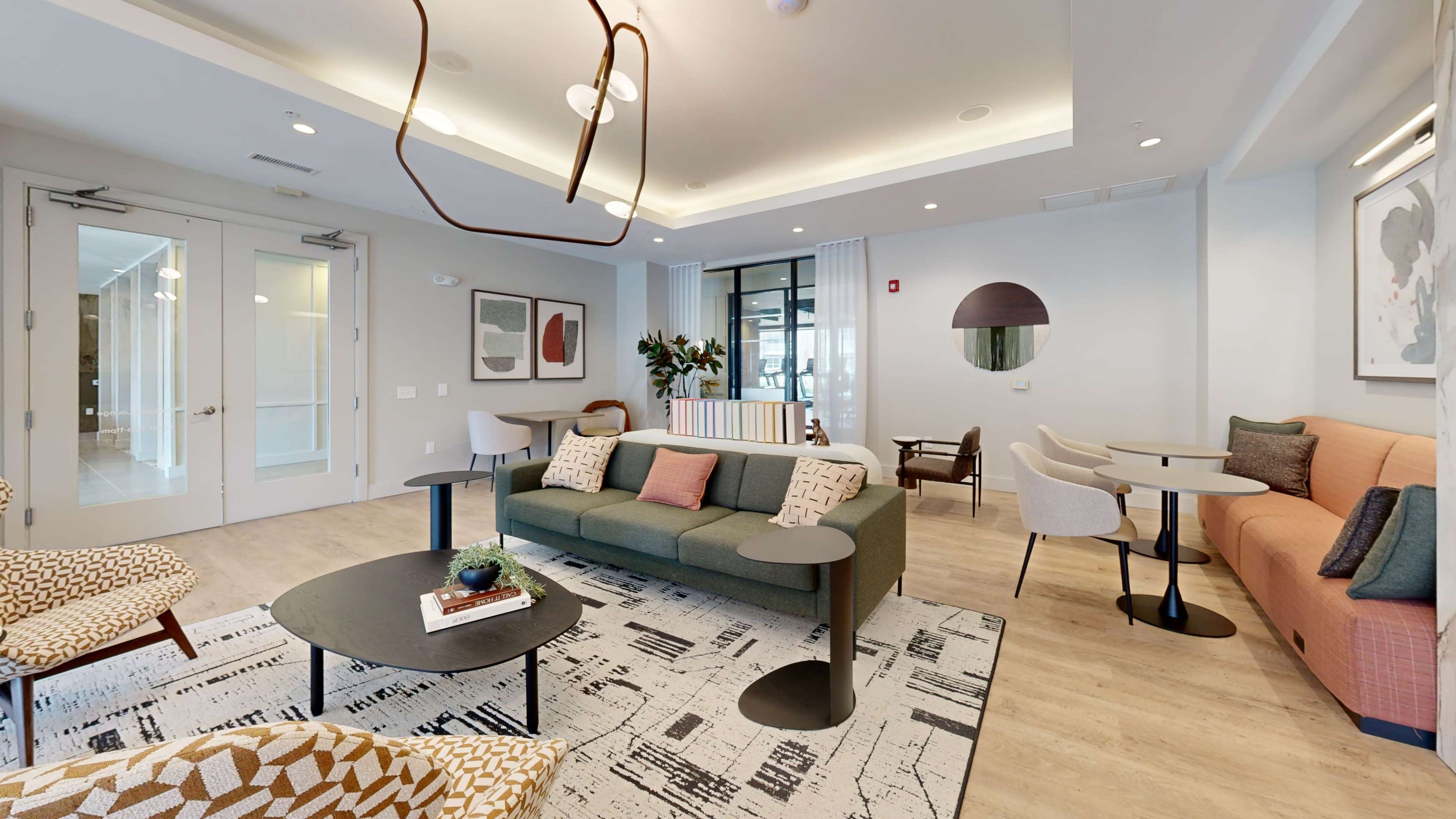 The image shows a modern lounge area featuring a green sofa, several chairs, and tables, with large windows and decorative artwork on the walls.